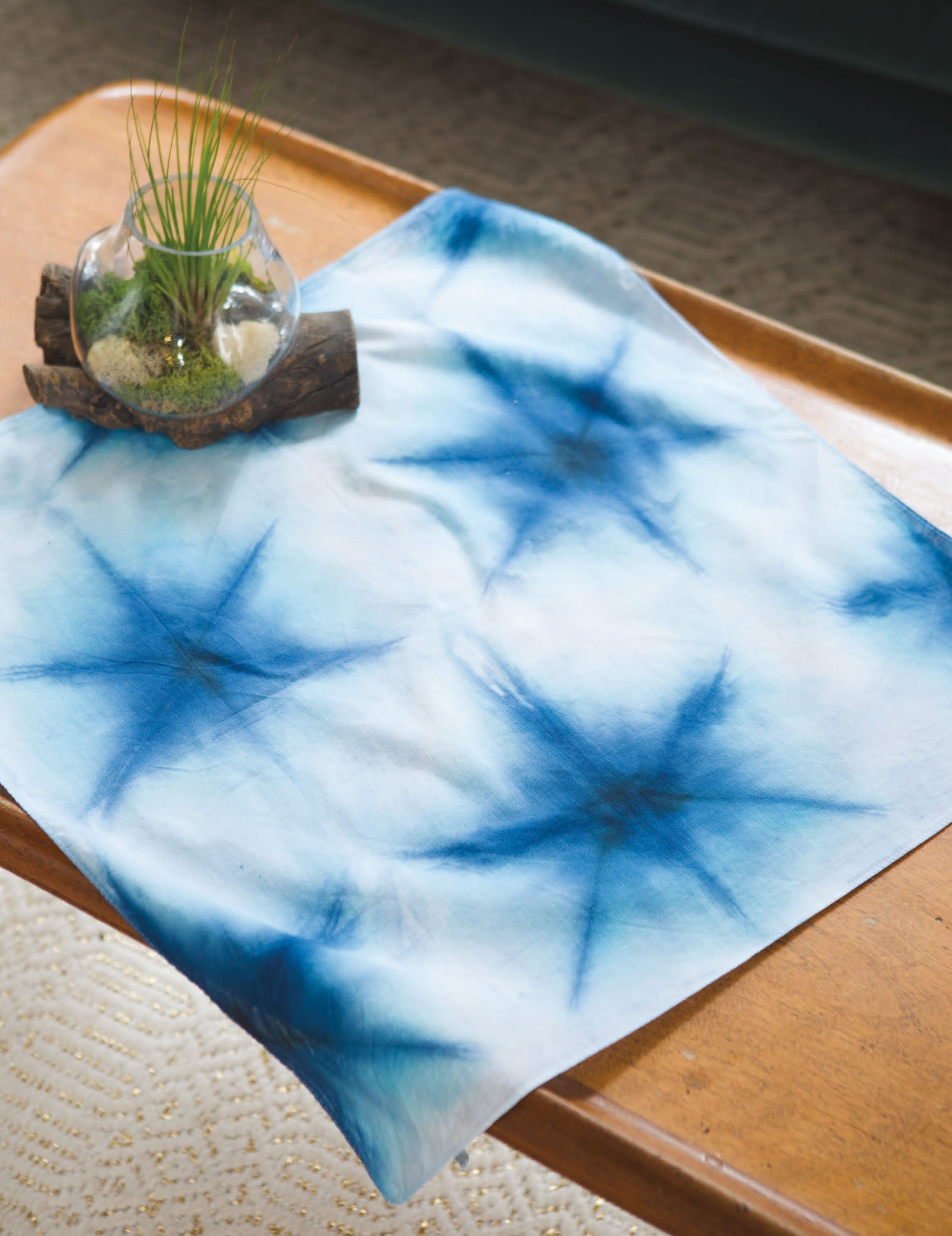 The "Shibori, Ice & Dip Dyeing" kit by Leisure Arts features a blue and white starburst-patterned cloth on a wooden table, with a glass bowl of plants, sand, and rocks displayed nearby atop driftwood. Perfect for beginner dyeing techniques.