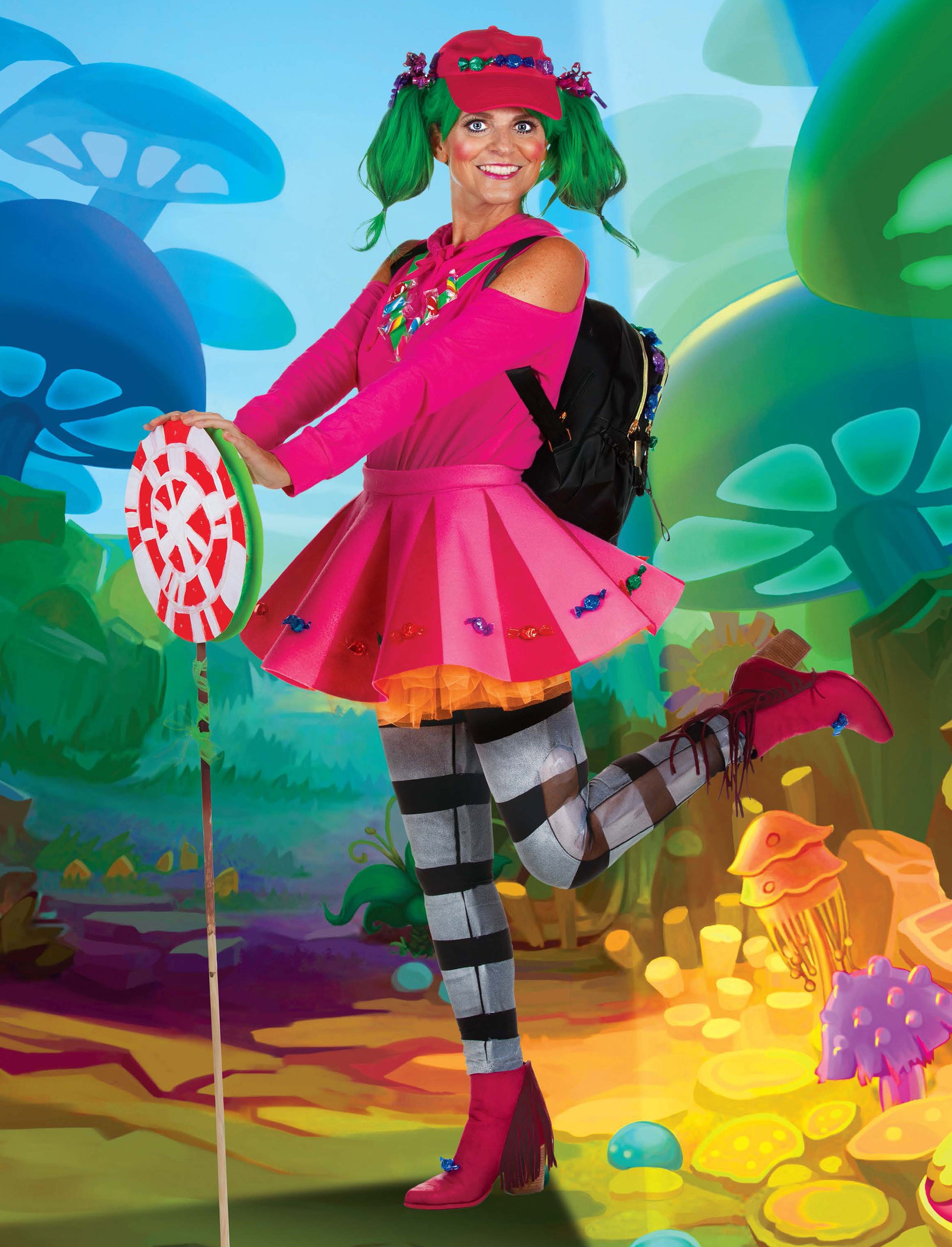 A woman dressed in a vibrant costume, made using low-sew techniques from the Cosplay DIY Workbook by Leisure Arts, stands on one leg in a fantasy forest, wearing a pink skirt, bright pink shoes, green pigtails, and holding a giant lollipop.