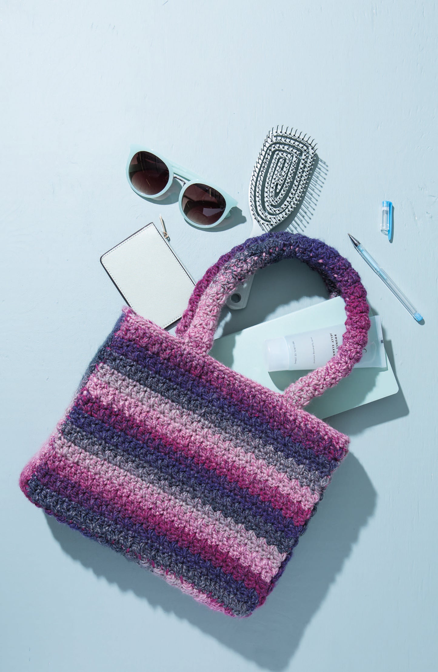 The Leisure Arts Crochet Handcrafted Handbags, featuring pink, purple, and gray stripes, sits on a light blue surface surrounded by sunglasses, a hairbrush, white wallet, blue book, paper, pen, and USB flash drive.