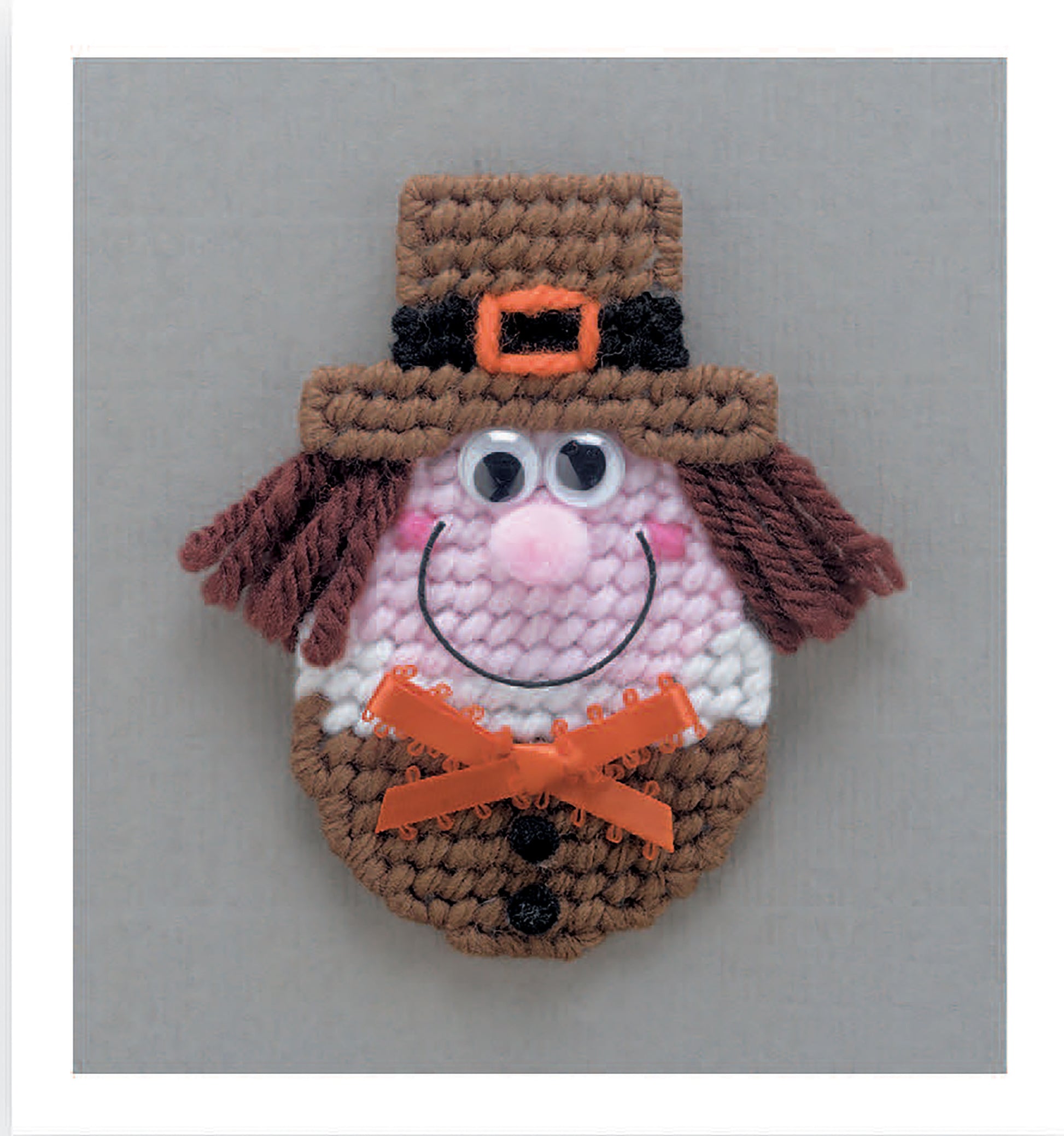 Leisure Arts' Magnet Board Creations in Plastic Canvas Digital Download features a beginner-friendly pilgrim face magnet with a brown hat, googly eyes, pink cheeks and nose, yarn hair, and orange bow—great for your fridge!.