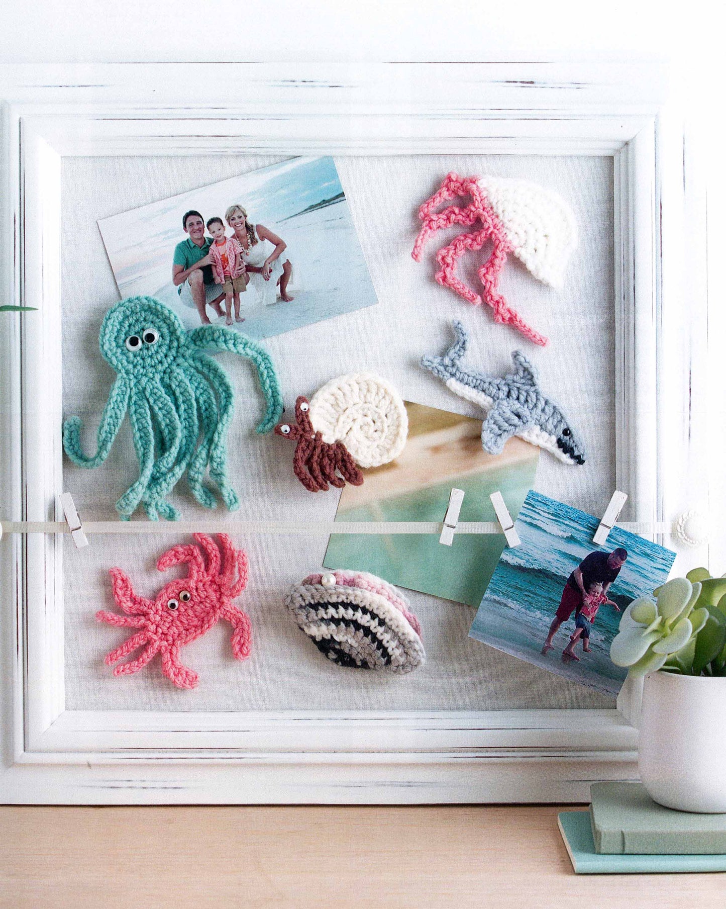 The Magnet Board Creations by Leisure Arts features a white-framed corkboard displaying two beach photos and six beginner crochet sea creatures—octopus, crab, jellyfish, nautilus, shark, and seashell—with a small succulent below.
