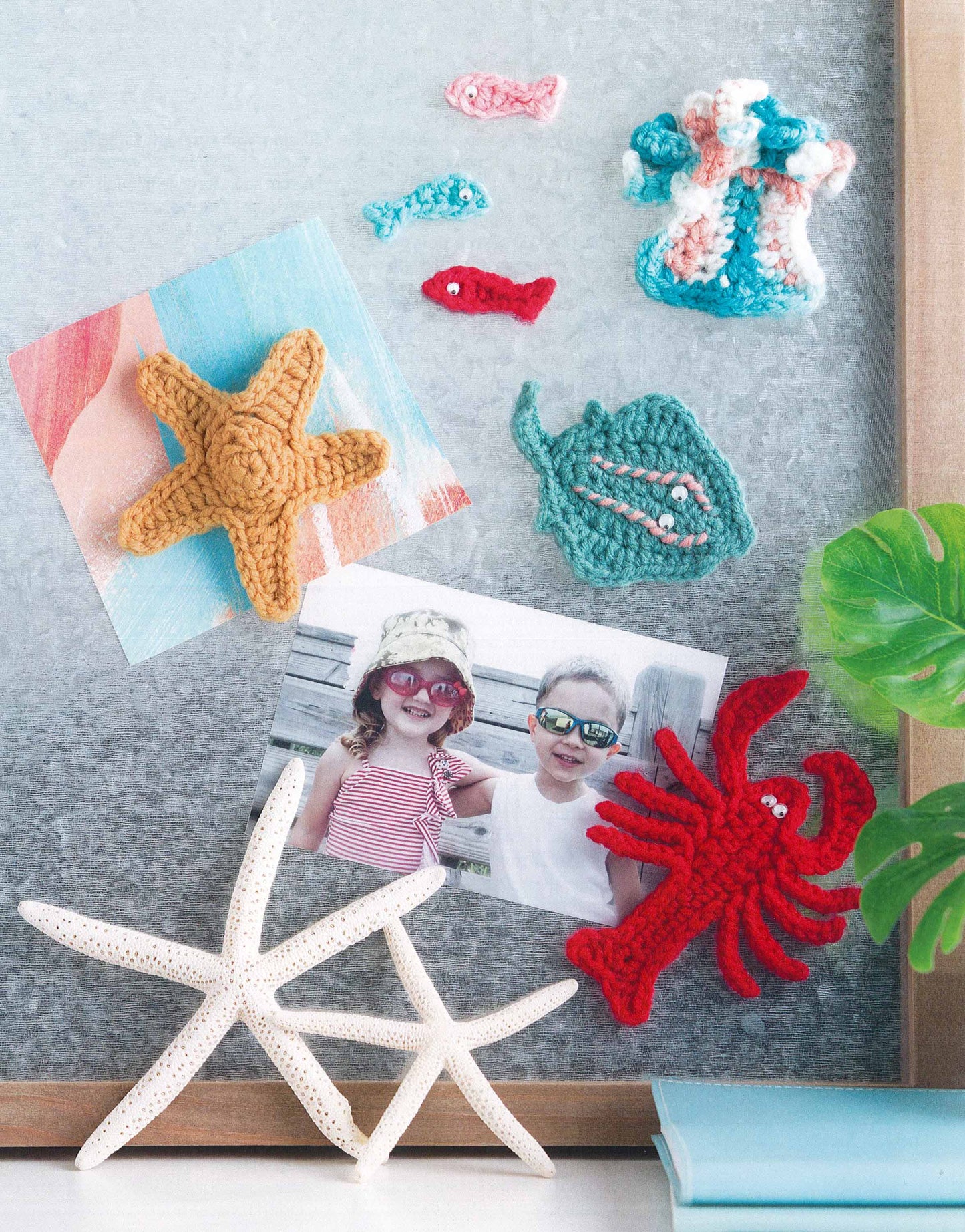 The Magnet Board Creations set by Leisure Arts features crocheted sea creatures—starfish, fish, stingray, lobster, and coral—from beginner patterns. Above them are photos of two kids in sun hats next to real starfish and a green plant.