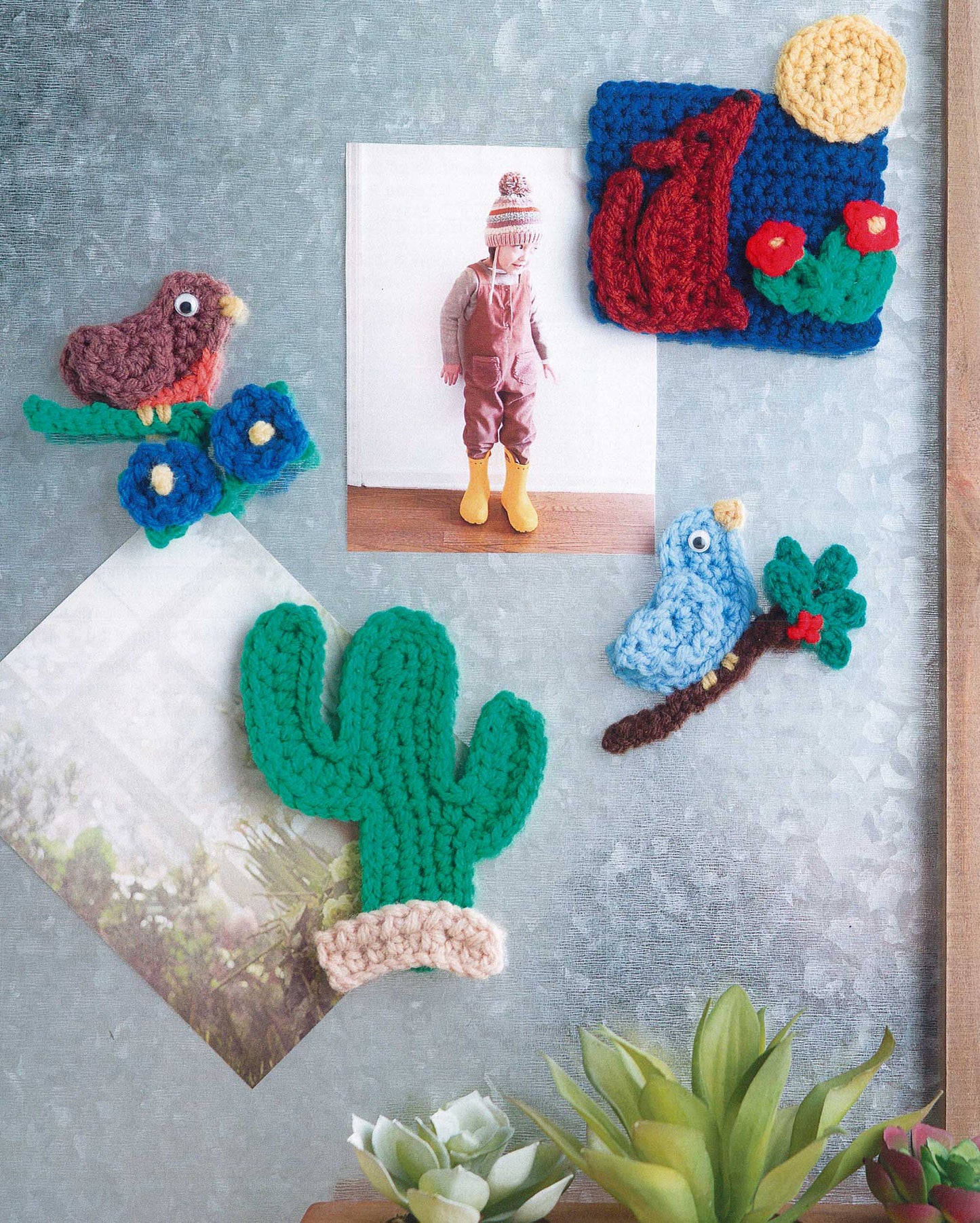 Magnet Board Creations by Leisure Arts displays two photos with crochet magnets shaped like a cactus, bird, blue branch, and landscape—fun accents for your fridge. A small succulent sits below for extra charm.