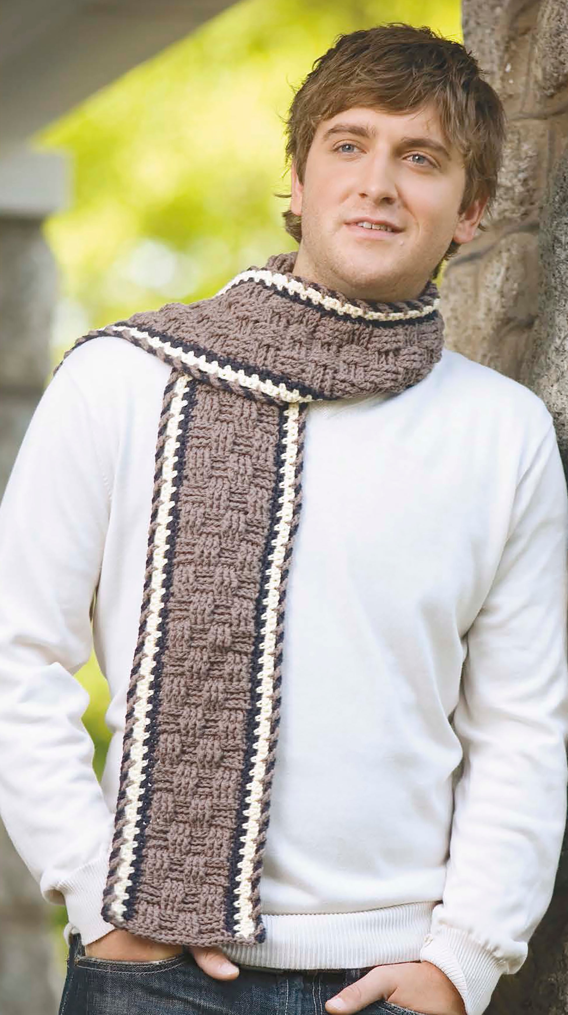 A man stands outdoors by a stone wall, relaxed and smiling slightly, wearing a white sweater and a brown crochet scarf with white and dark borders from "The Little Encyclopedia of Crocheted Scarves" by Leisure Arts.