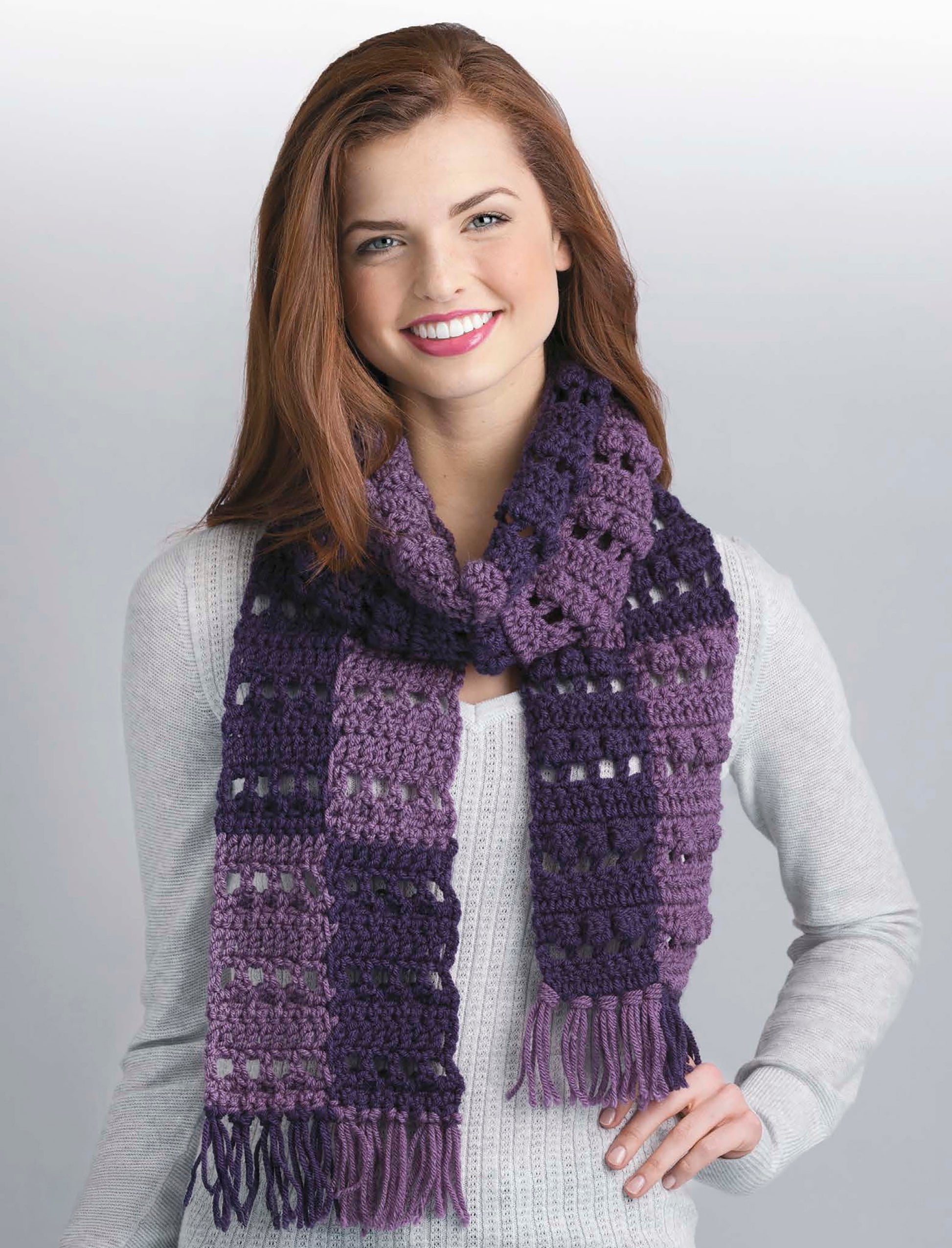 A smiling woman wears a light gray sweater and chunky purple scarf with fringe, standing against a plain background—inspiring you to try 20 designs from The Little Encyclopedia of Crocheted Scarves by Leisure Arts or explore expert scarf styling tips.