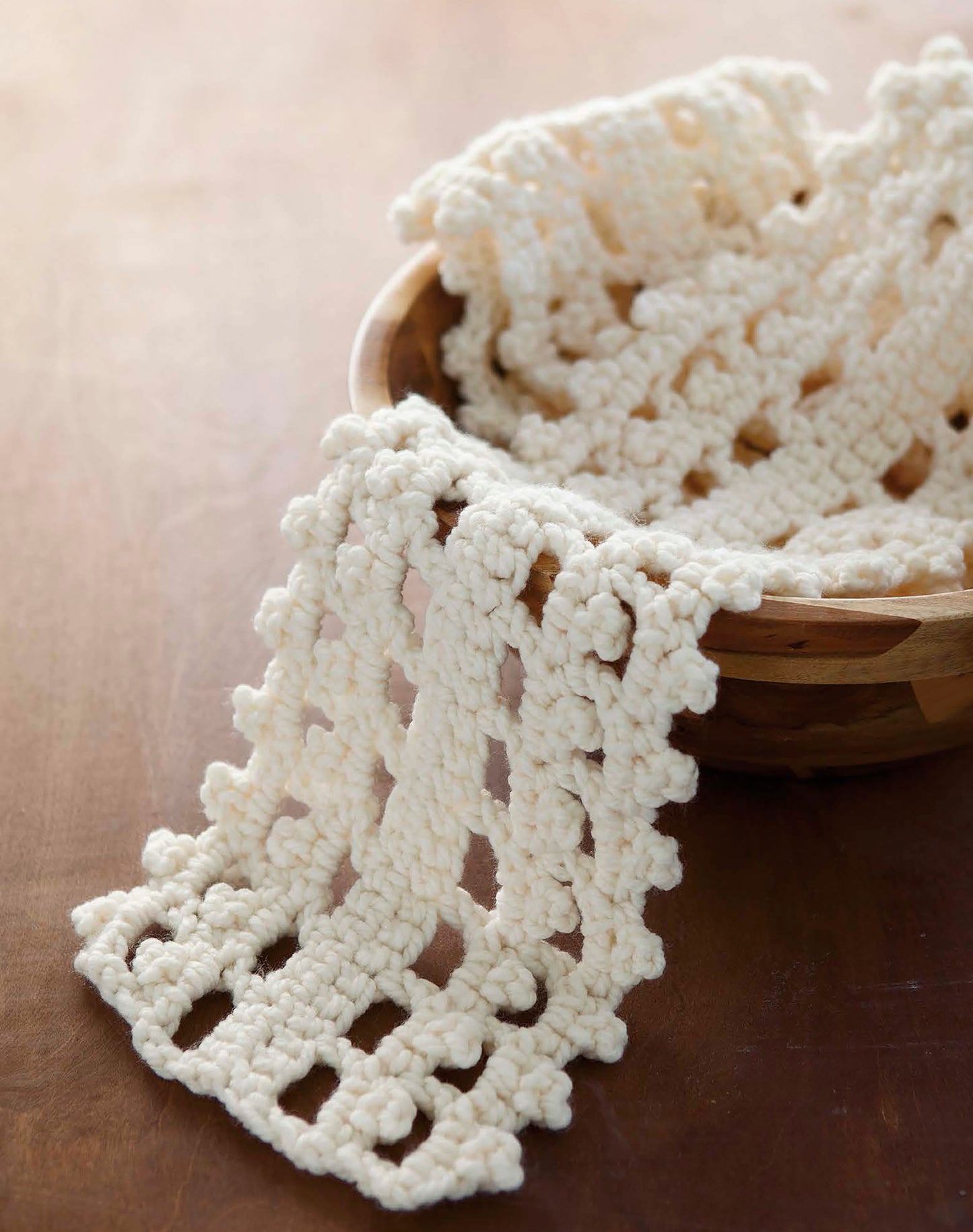 A cream hand-crocheted scarf with a lacy, openwork pattern from "The Little Encyclopedia of Crocheted Scarves" by Leisure Arts drapes out of a wooden bowl onto a brown surface.