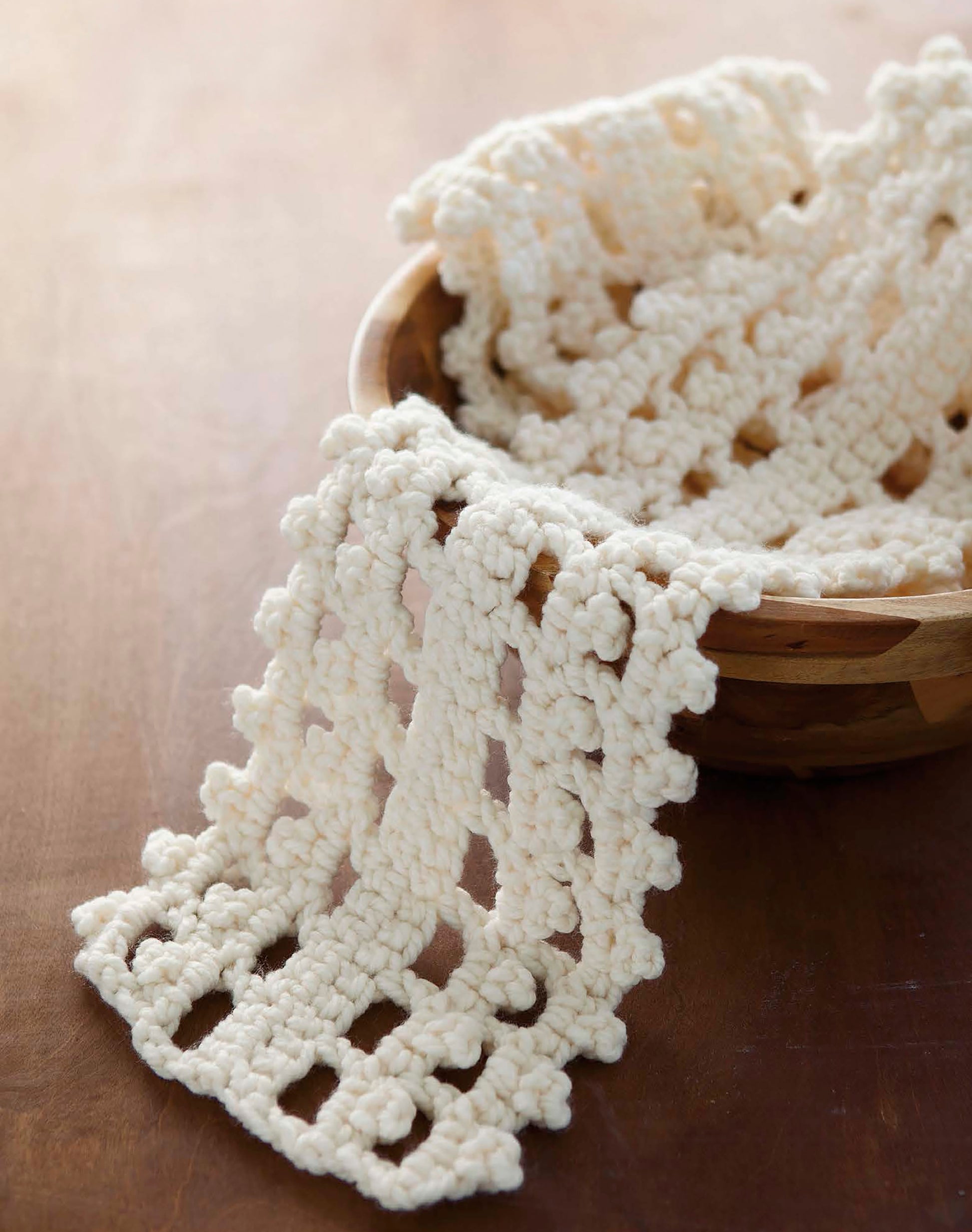 A cream hand-crocheted scarf with a lacy, openwork pattern from "The Little Encyclopedia of Crocheted Scarves" by Leisure Arts drapes out of a wooden bowl onto a brown surface.