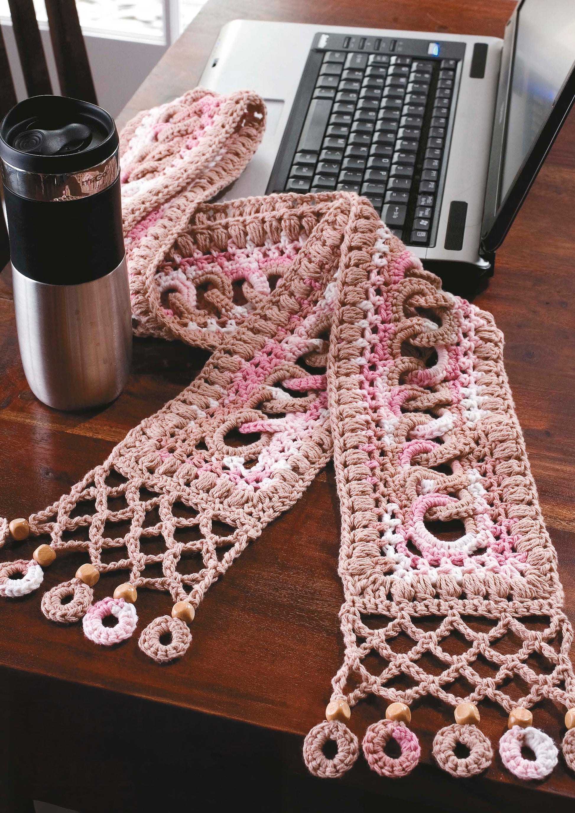 The Little Encyclopedia of Crocheted Scarves by Leisure Arts rests on a wooden table near a laptop and travel mug, showcasing a pink and beige scarf with loops and beads, plus 20 creative designs and expert crochet tips.
