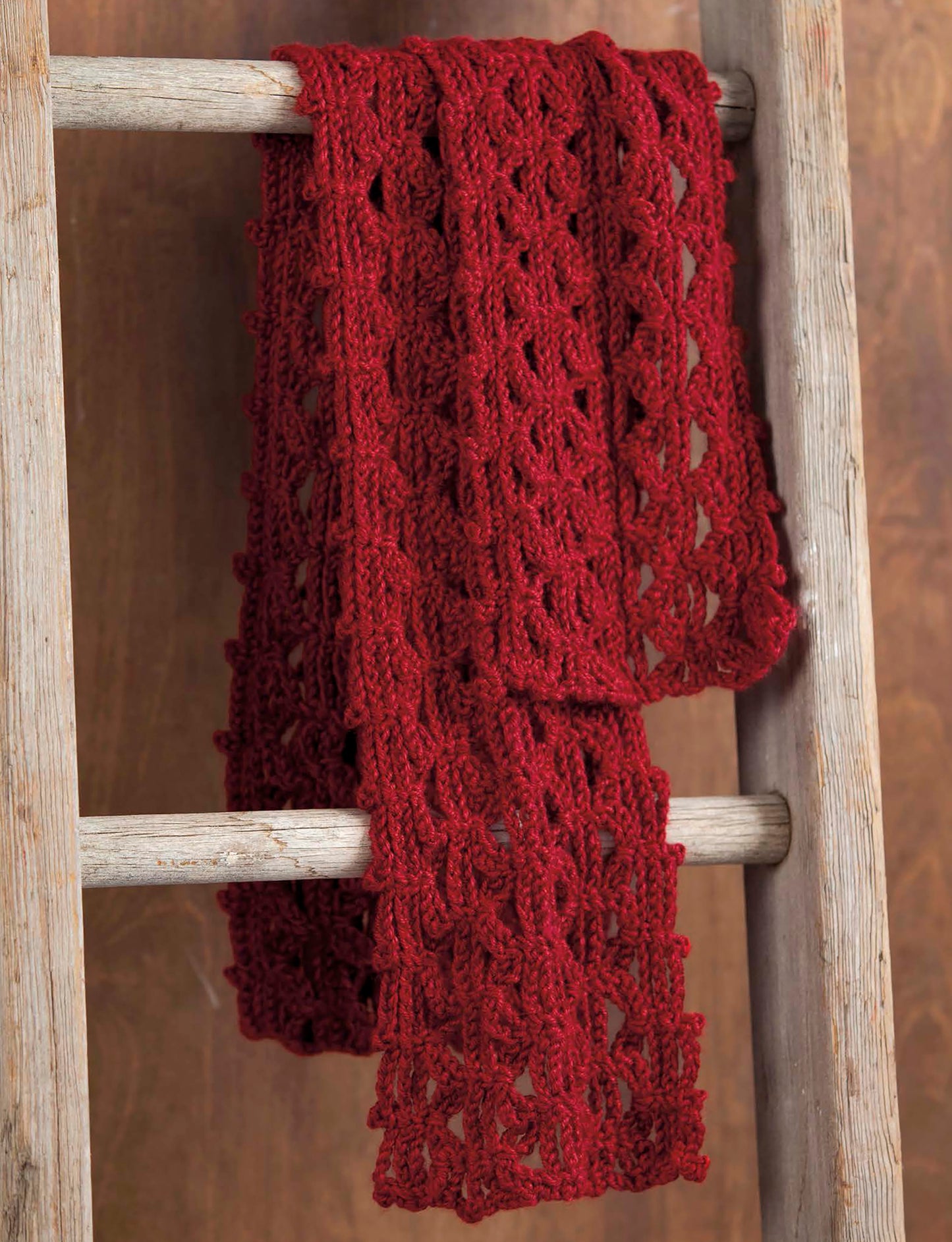 A red, lacy crochet scarf from The Little Encyclopedia of Crocheted Scarves by Leisure Arts is draped over a rustic wooden ladder against a brown background.