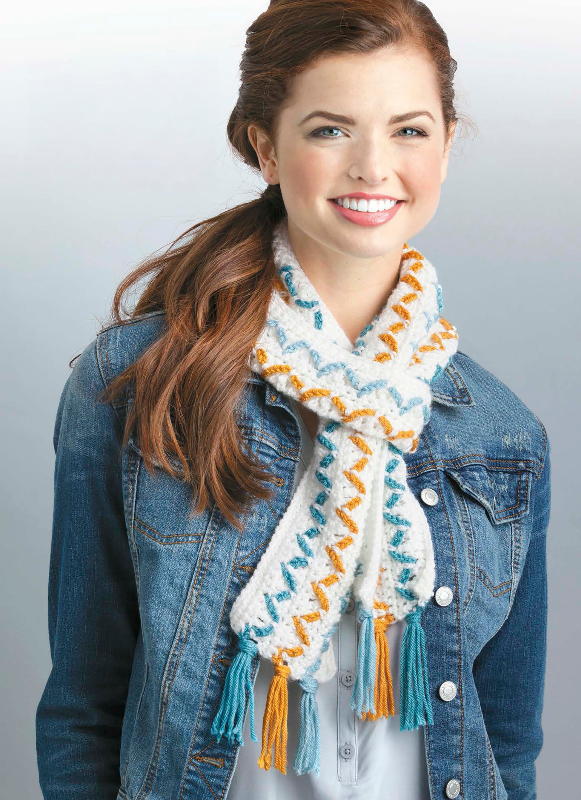 A smiling woman with long brown hair in a ponytail wears a blue denim jacket over a white shirt and a Leisure Arts scarf from The Little Encyclopedia of Crocheted Scarves, featuring expert tips and blue-orange zigzag patterns inspired by 20 designs.