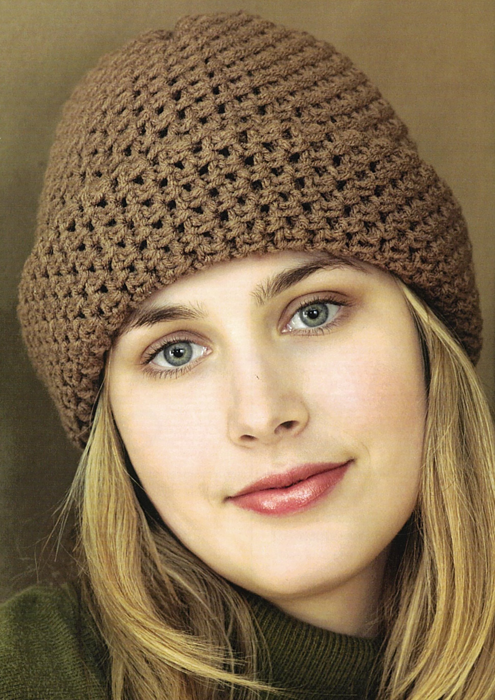 A woman with light skin and blonde hair wears a brown crocheted beanie made using Leisure Arts’ Crochet Made Easy, paired with a green sweater, smiling gently at the camera against a plain background.