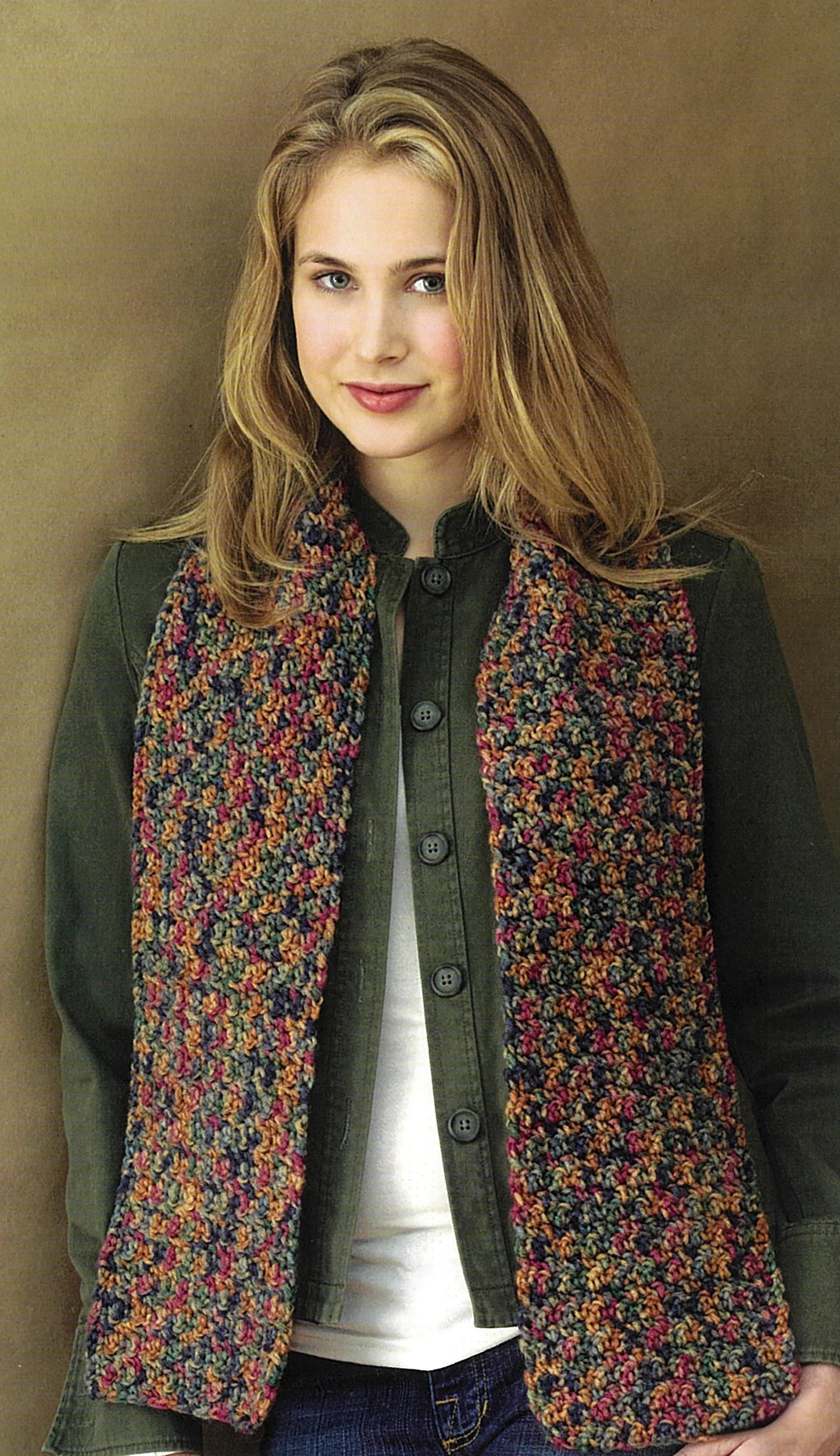 A woman with long blonde hair wears a dark green jacket, white shirt, and a colorful chunky knit scarf from Leisure Arts' "Crochet Made Easy"—ideal for beginners wanting to try 8 projects. She stands against a brown background, smiling softly at the camera.