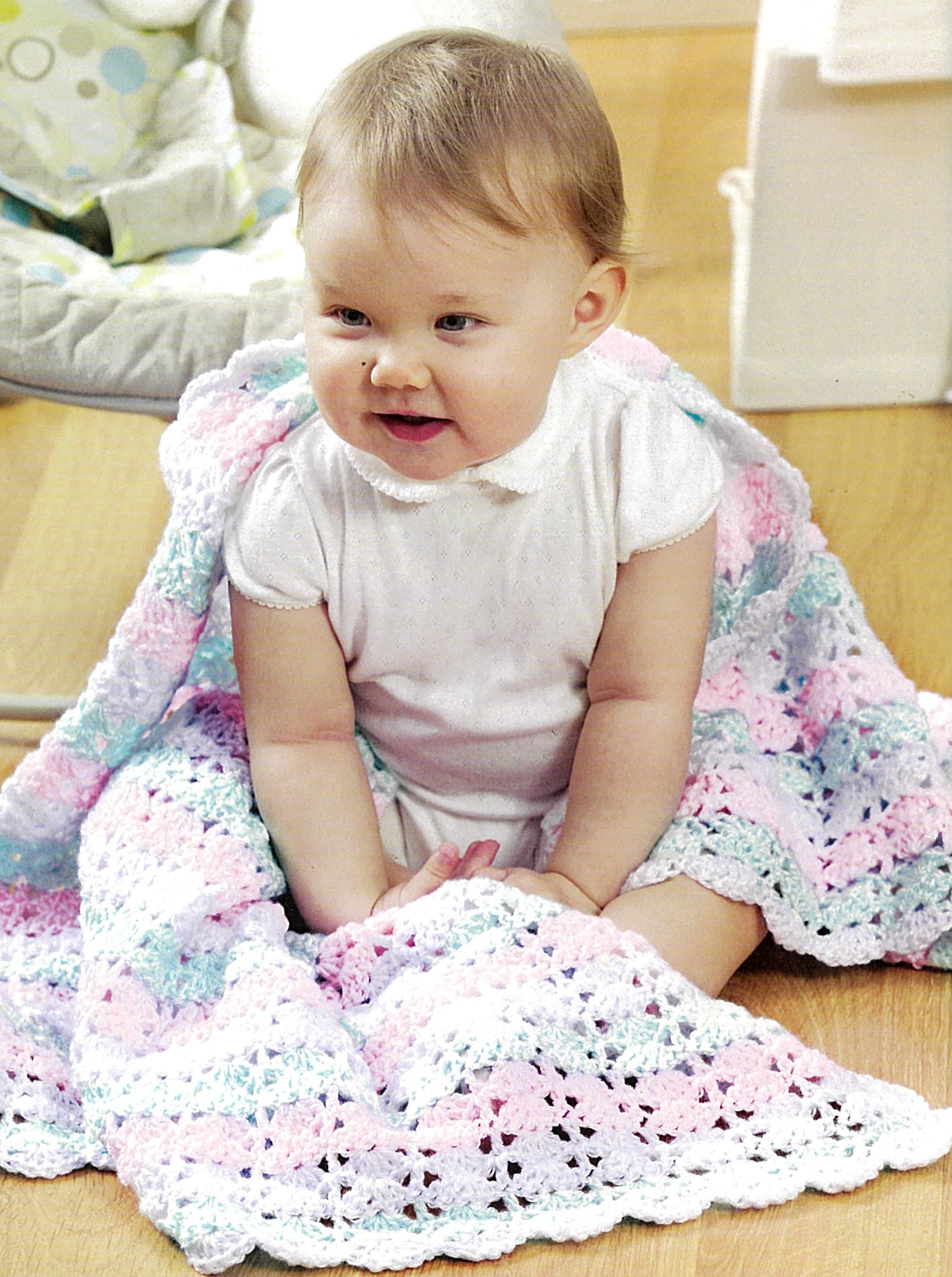 A baby in white sits on a wooden floor, wrapped in a pastel crocheted blanket made with Crochet Made Easy by Leisure Arts, a beginner-friendly collection featuring 8 projects. The baby looks up with a cheerful expression.