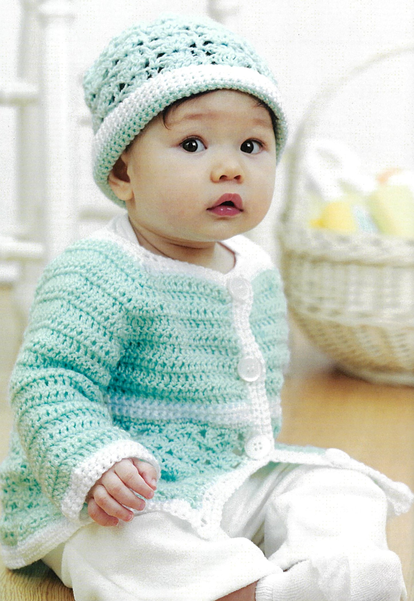 A baby wears a light blue crocheted hat and matching cardigan with white trim and buttons. Inspired by Leisure Arts’ “Crochet Made Easy,” this charming look is one of 8 beginner-friendly projects. A white basket with pastel items sits blurred in back.
