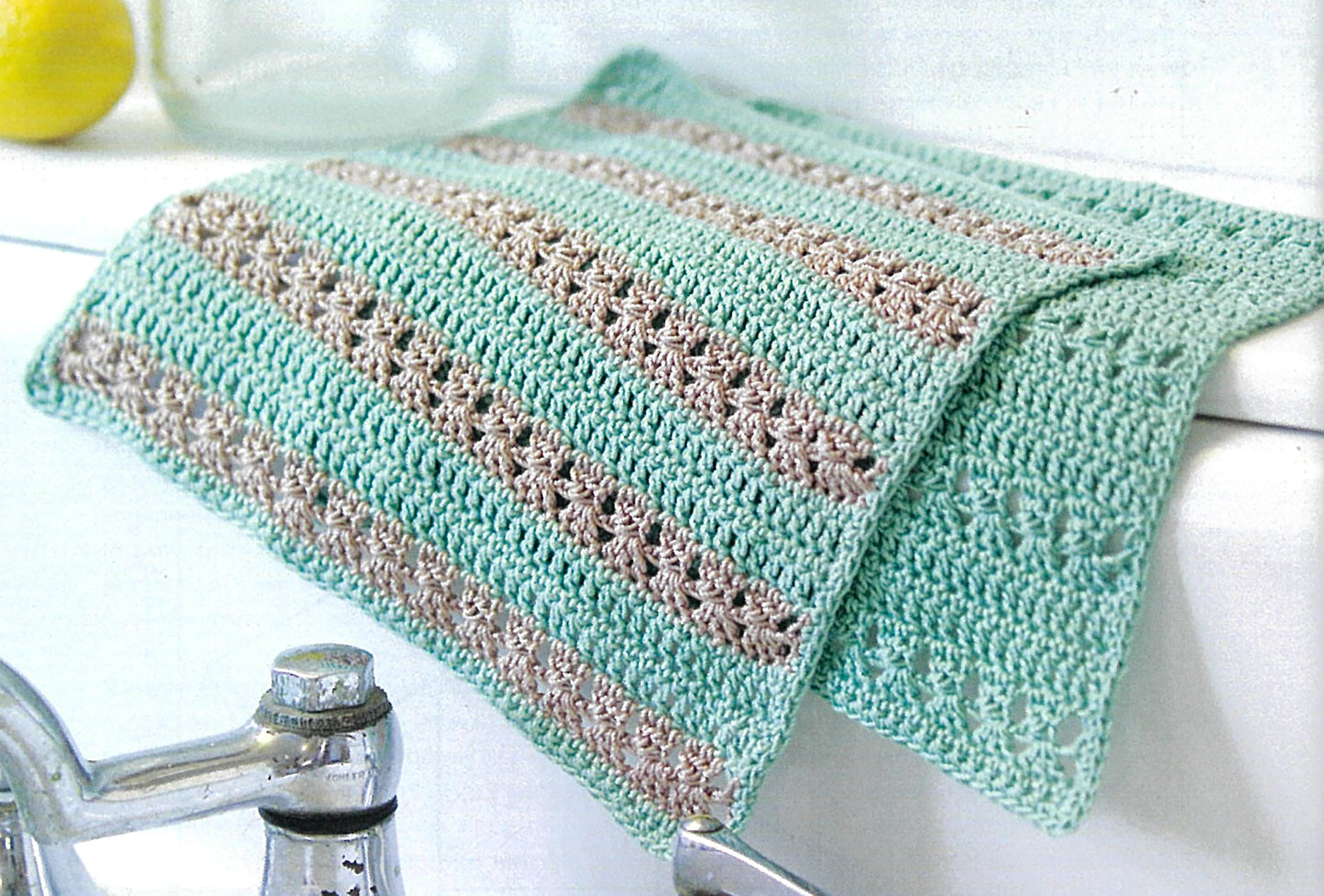 The Crochet Made Easy cloth by Leisure Arts features alternating mint green and beige stripes with openwork details, displayed draped over a white sink beside a silver faucet—perfect for beginners.