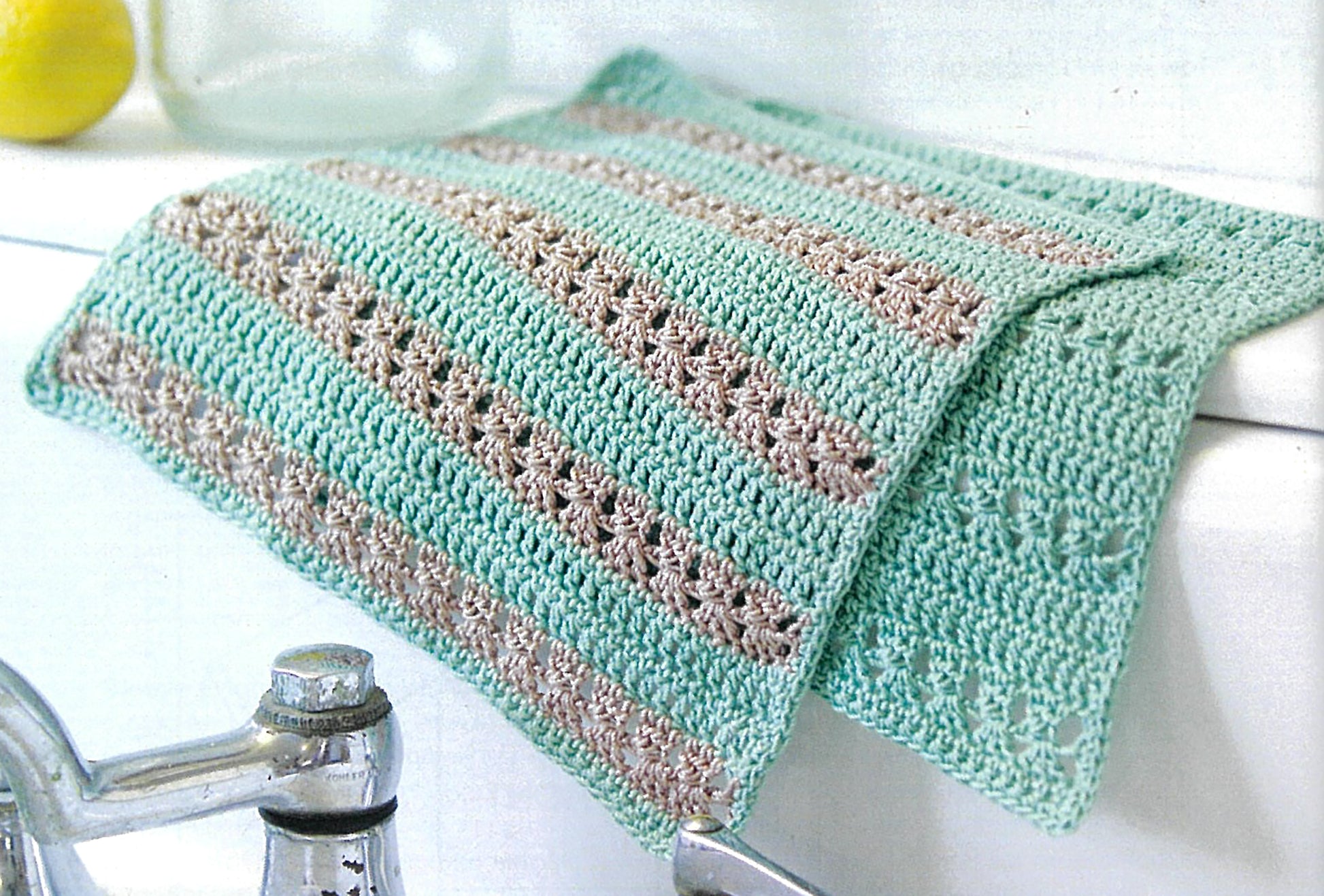The Crochet Made Easy cloth by Leisure Arts features alternating mint green and beige stripes with openwork details, displayed draped over a white sink beside a silver faucet—perfect for beginners.