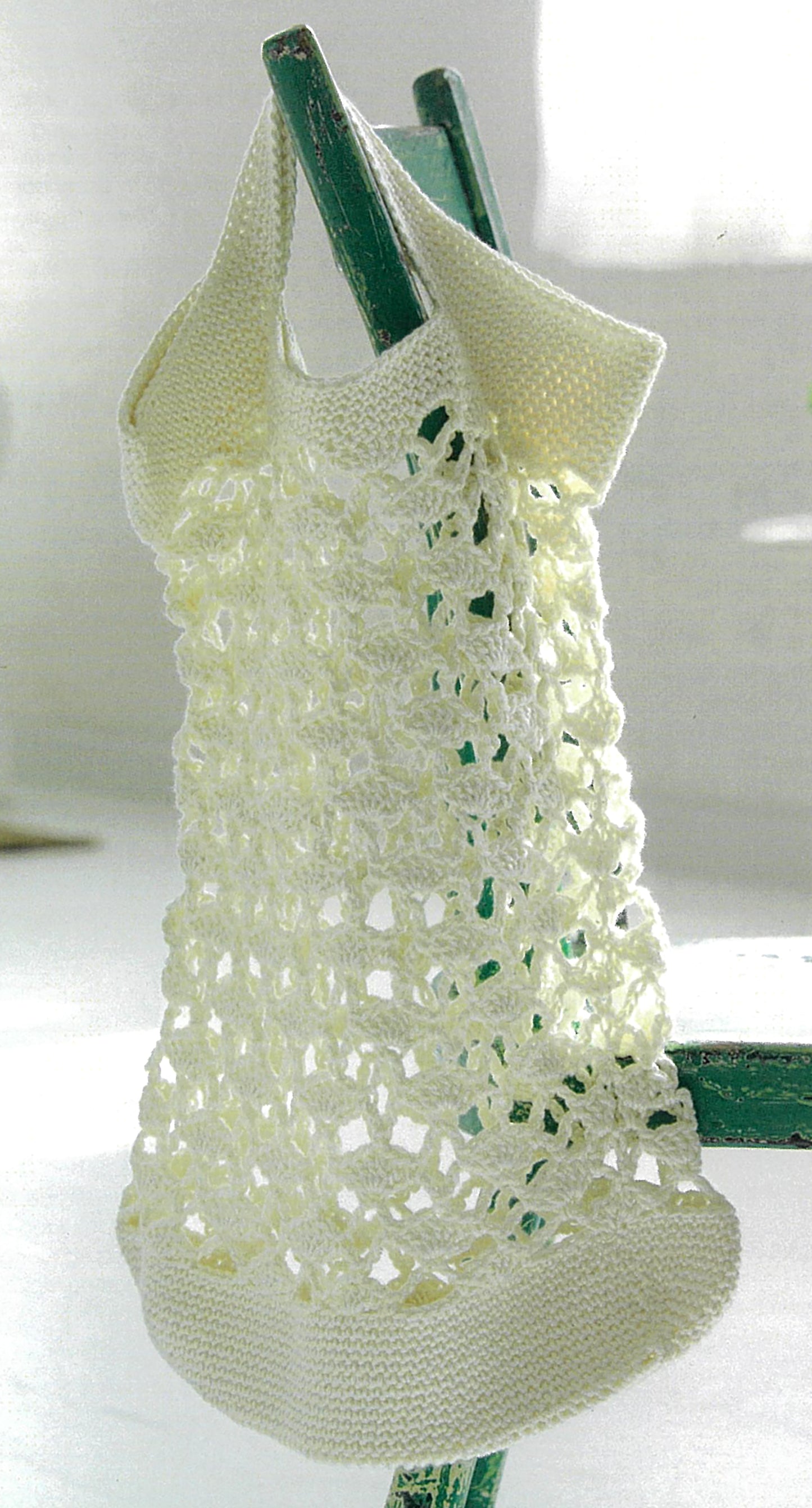 A handmade white open-weave knit tote, perfect for beginners, hangs on a green chair in a sunlit room; inspired by Leisure Arts' Crochet Made Easy and its 8 creative afternoon projects.