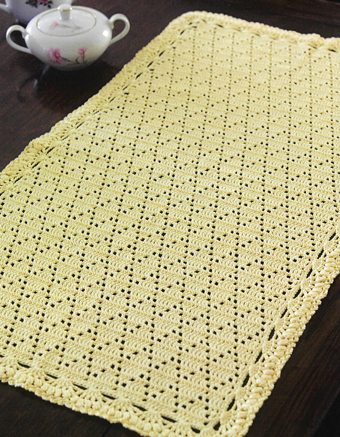 A rectangular, off-white crocheted table mat with a textured pattern and decorative border from Leisure Arts’ Crochet Made Easy rests on a dark wooden table beside a white floral teapot—ideal for beginner crafters.