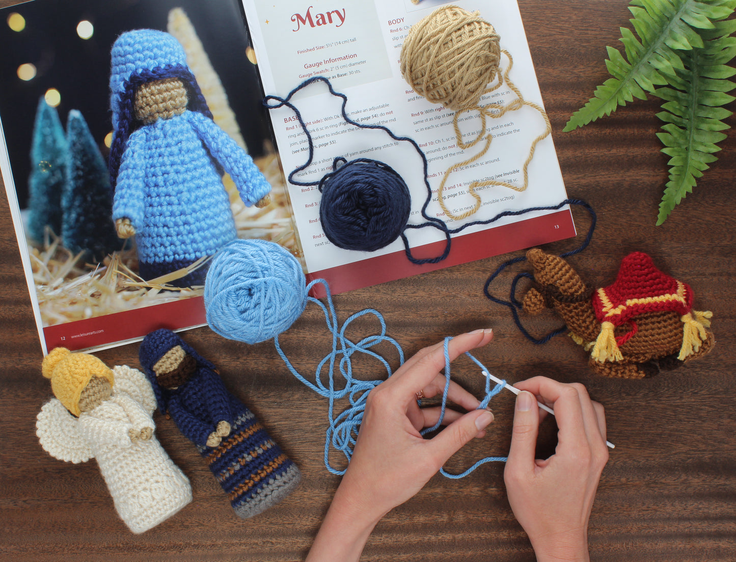 Hands crochet with blue yarn next to yarn balls, crafting beginner amigurumi nativity figures using the Crochet Nativity Digital Download by Leisure Arts, with an open pattern book and a green plant on a wooden table.