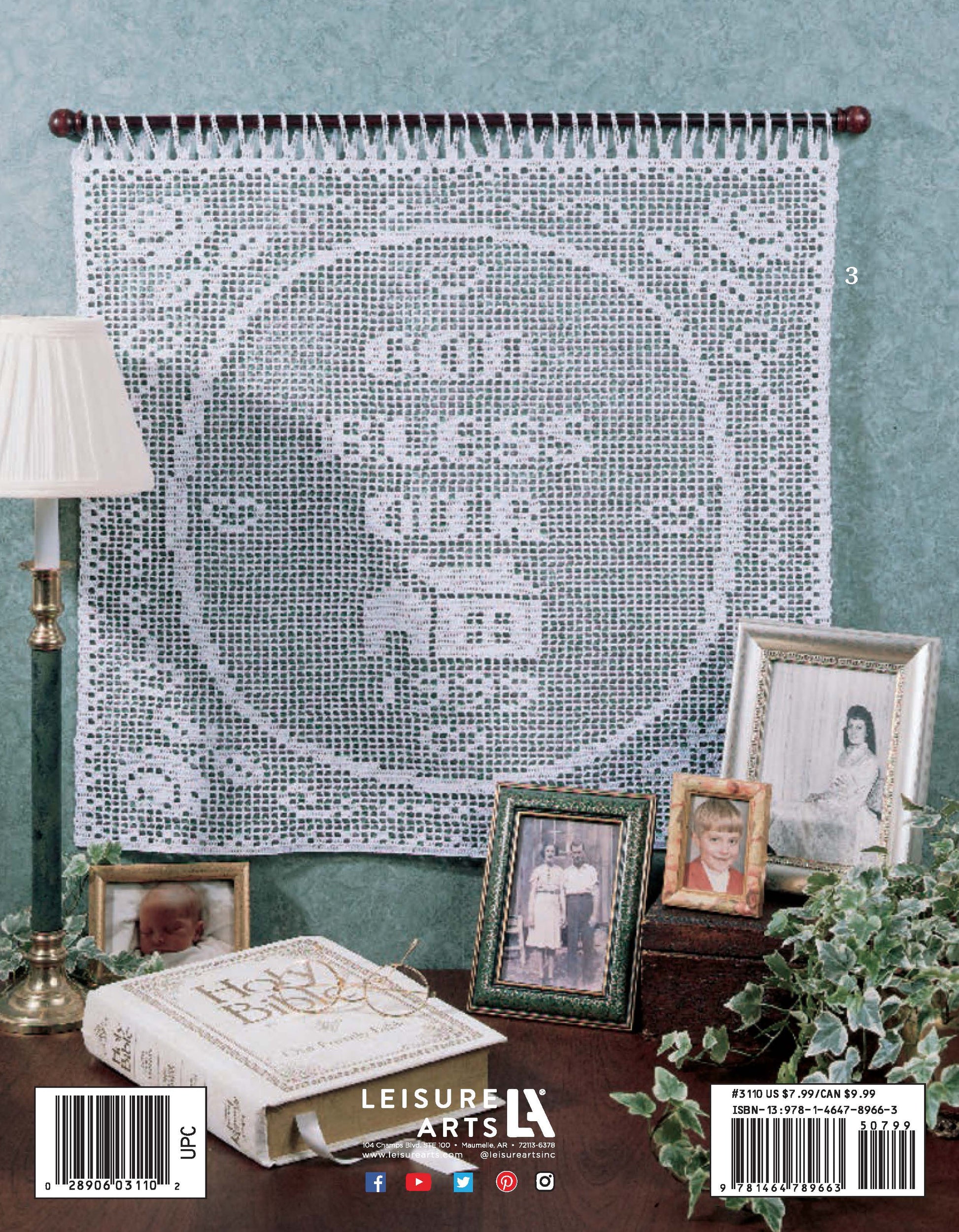 A crocheted wall hanging made with the “Your Name & More in Filet Crochet” pattern from Leisure Arts, featuring an elegant initial motif and the words “God Bless Our Home,” hangs behind framed family photos, a lamp, and an open Bible on a wooden table.