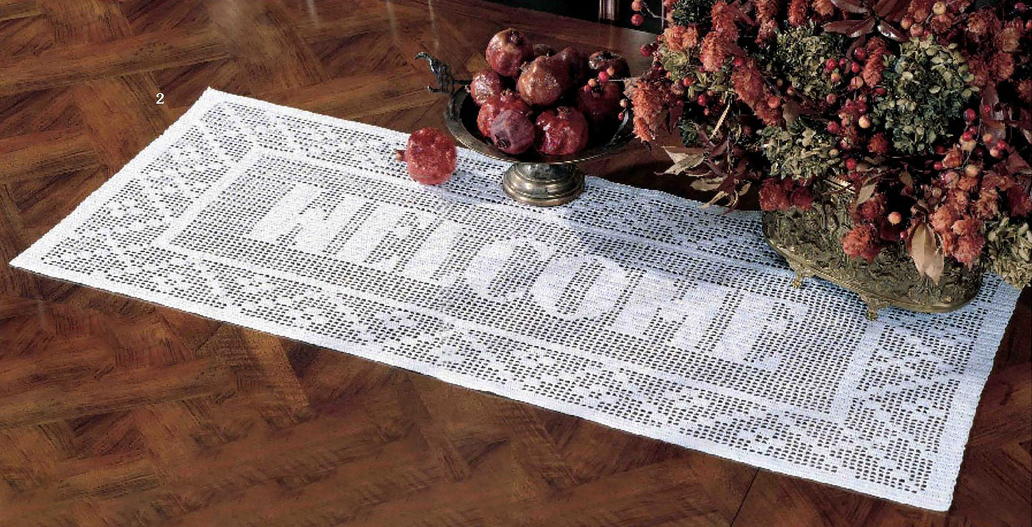 A white crochet table runner from Leisure Arts’ “Your Name & More in Filet Crochet” collection, featuring an elegant initial motif and the word WELCOME, is displayed on a wooden table adorned with a bowl of red fruit and a bouquet of dried flowers.