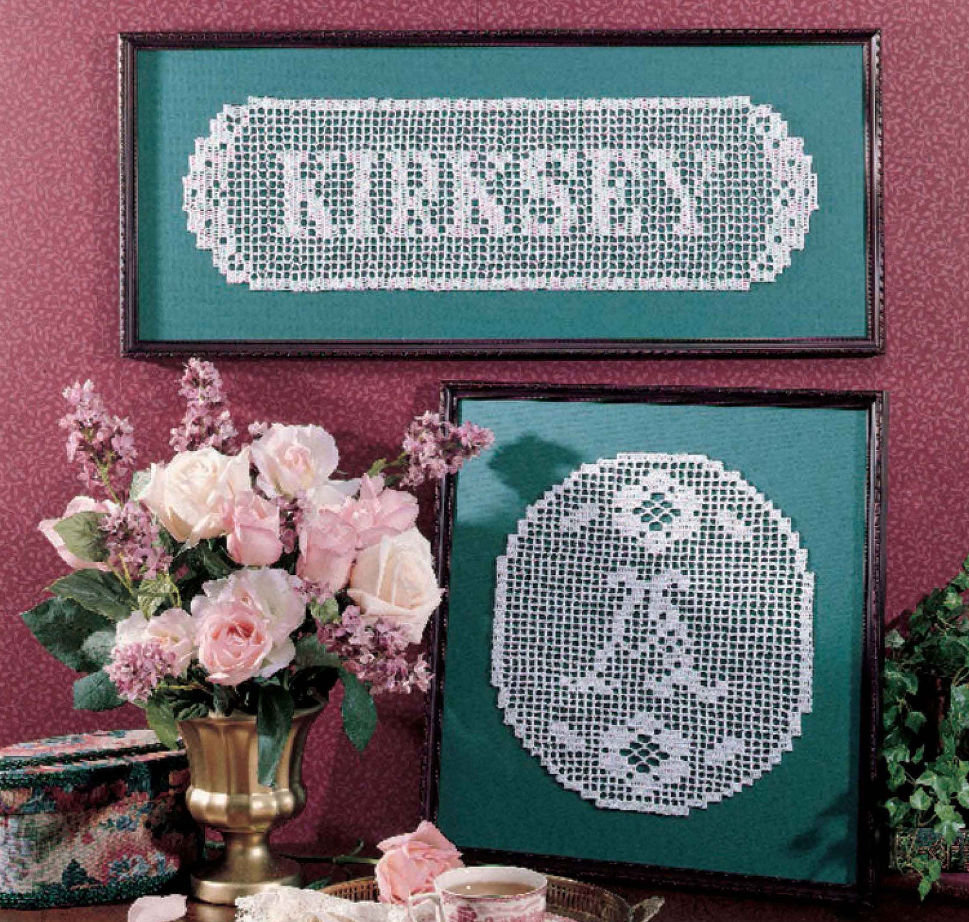 Two framed laceworks, created using the Leisure Arts “Your Name & More in Filet Crochet” digital download pattern, showcase bedspread weight cotton yarn artistry: the top piece displays a family name crochet “KIRKSEY” in white atop green fabric, while the bottom features a monogram “A” surrounded by delicate floral filet crochet motifs. Both are beautifully arranged near a vase of pink roses on a table.