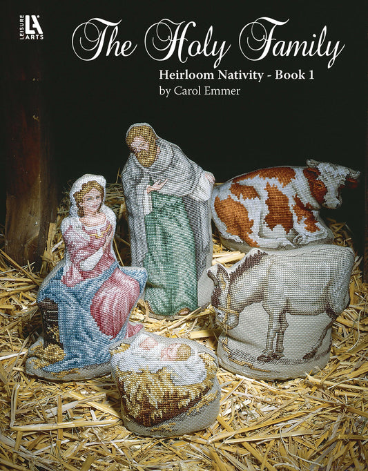 The Holy Family Heirloom Nativity Book 1 by Leisure Arts features a cross-stitch nativity scene of Mary, Joseph, and baby Jesus on straw with a cow and donkey nearby.