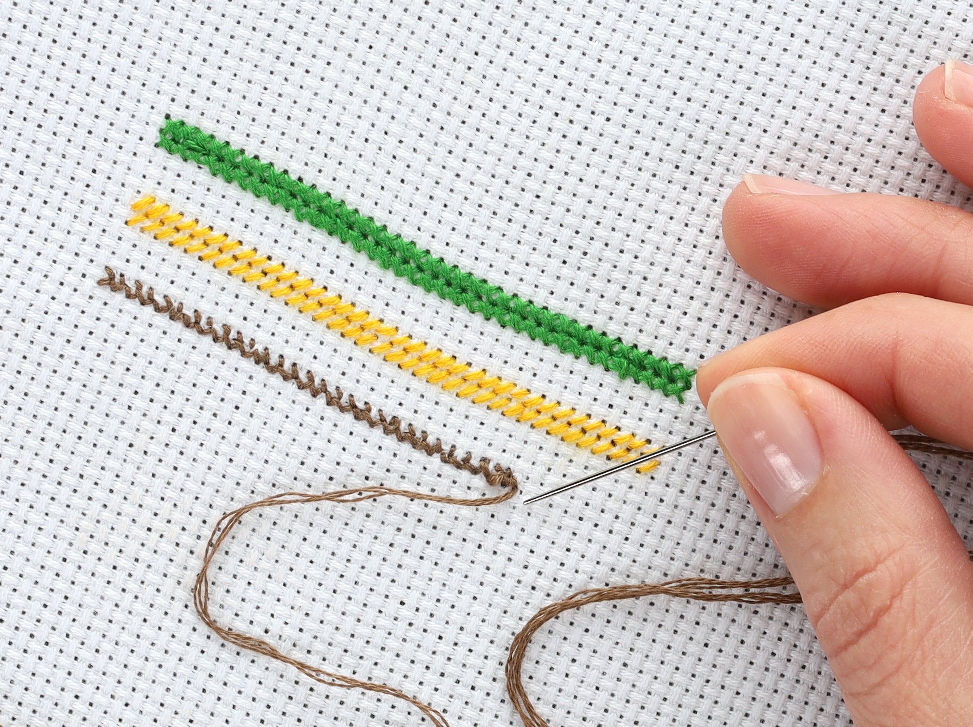 A hand stitches brown thread onto white cross stitch fabric beside finished green, yellow, and brown lines—ideal for Leisure Arts projects like The Shepherds Heirloom Nativity Book 3.
