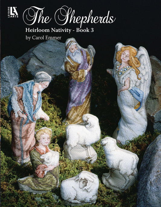 The Shepherds Heirloom Nativity Book 3 by Leisure Arts features cross-stitched nativity figurines—shepherds, sheep, and an angel—artfully displayed on moss with rocks, showcasing beautiful holiday designs.
