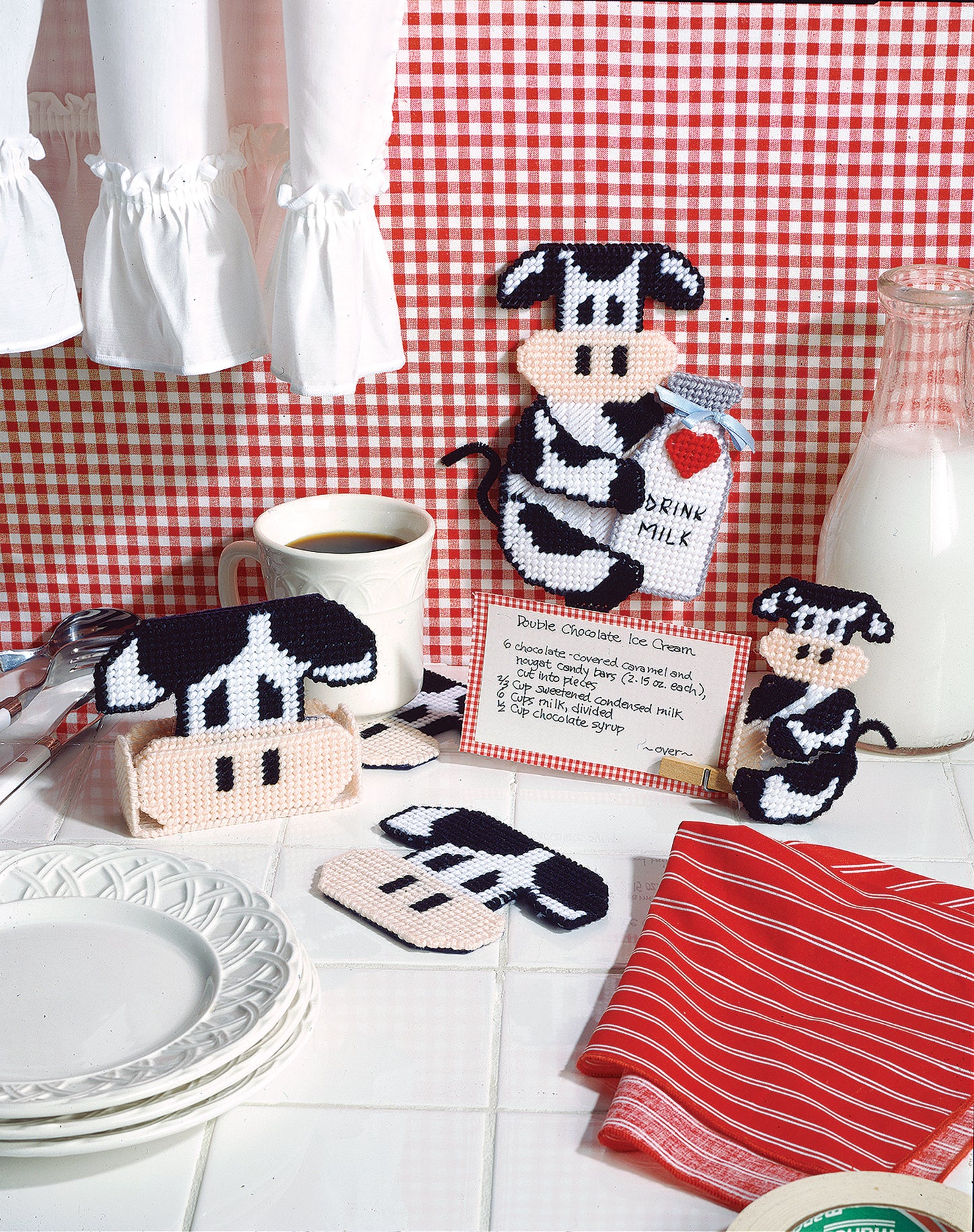 A cozy kitchen setting features Leisure Arts’ Barnyard Buddies in Plastic Canvas, perfect for beginners, alongside a glass milk bottle, coffee cup, white plates, and a red-striped towel on a red checkered tablecloth.