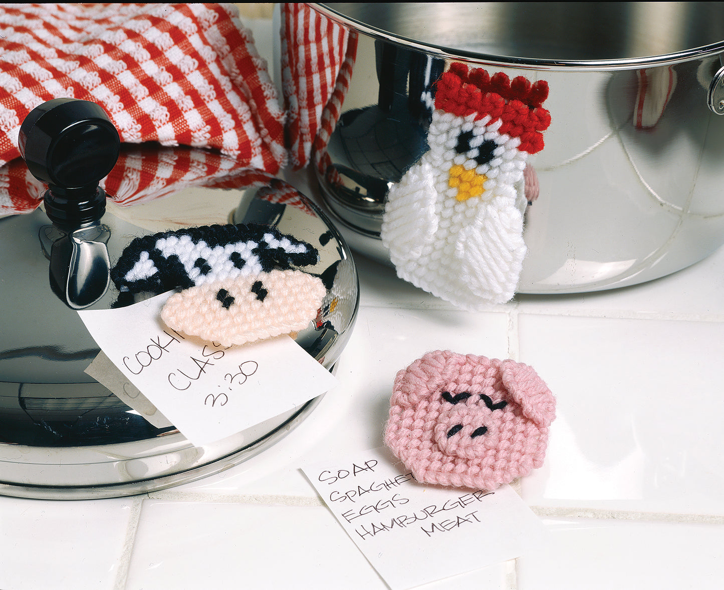 Leisure Arts’ Barnyard Buddies in Plastic Canvas features cow, chicken, and pig magnets on shiny metal pots. A red and white towel and handwritten notes rest on a white tiled surface—perfect for your next kitchen project.