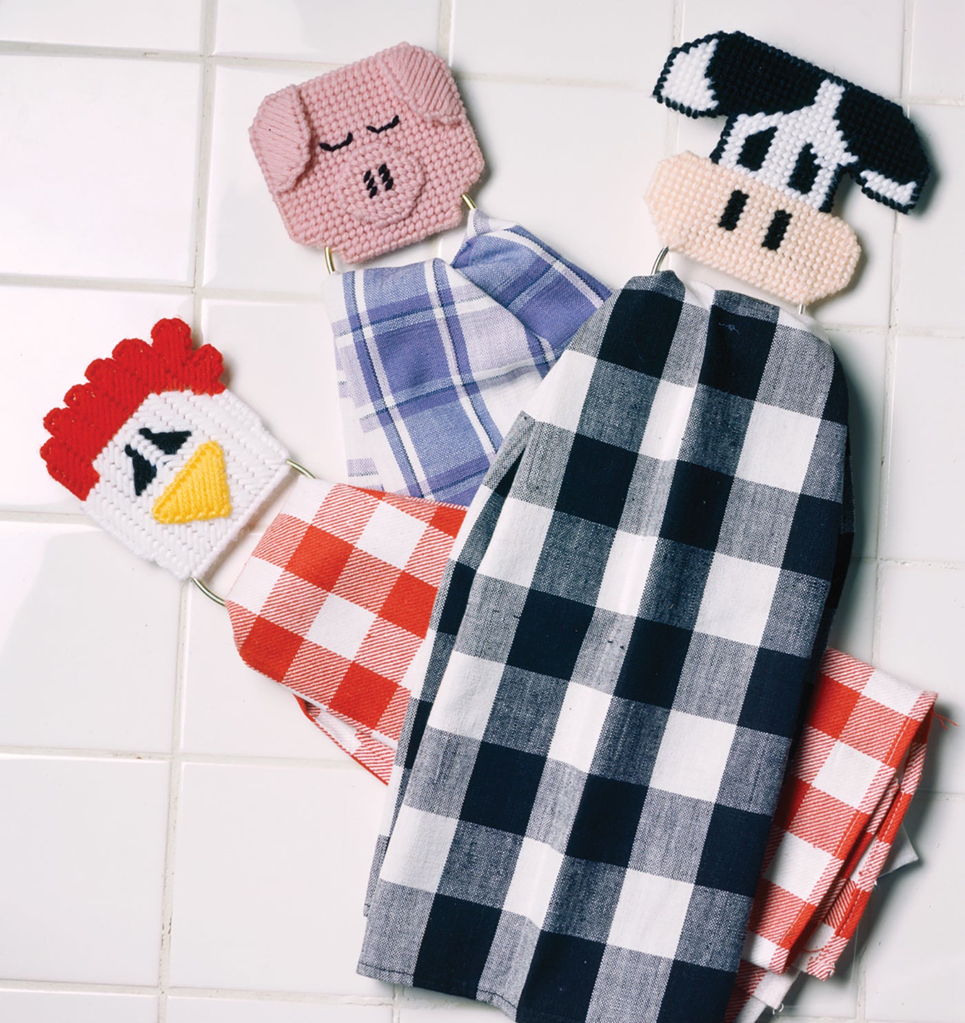 Three checkered dish towels are displayed on a white tiled surface, each held by a beaded animal face holder—pig, cow, and chicken—from the "Barnyard Buddies in Plastic Canvas" kit by Leisure Arts for beginner kitchen stitching projects.
