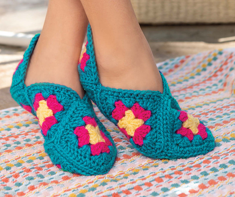 Close-up of feet in Leisure Arts' Great Grannies Made Modern slippers—blue base with pink and yellow granny squares—resting on colorful woven fabric.
