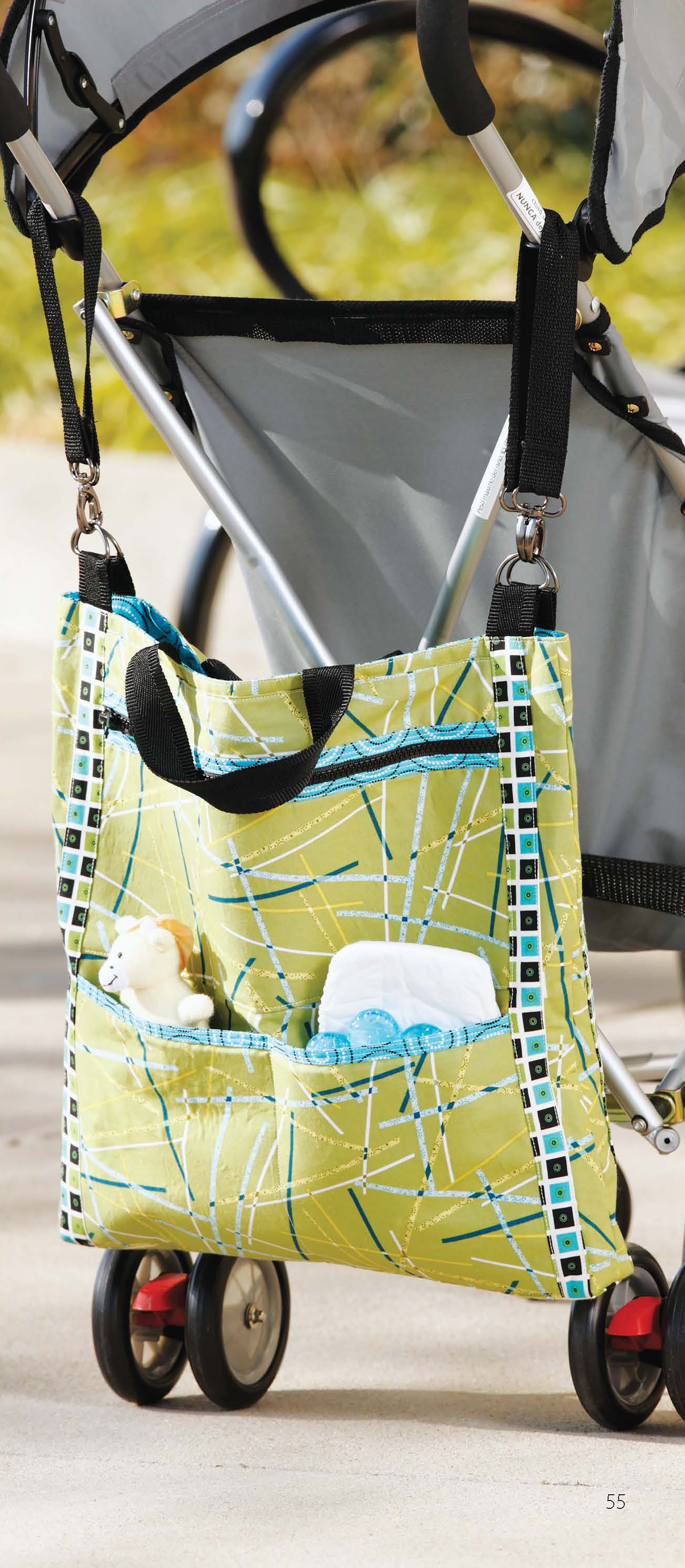 The “It’s in the Bag! Digital Download” by Leisure Arts features a green patterned diaper bag with black handles, made from beginner sewing projects. Hanging on a stroller outdoors, its front pocket holds a stuffed animal, white object, and blue cloth.