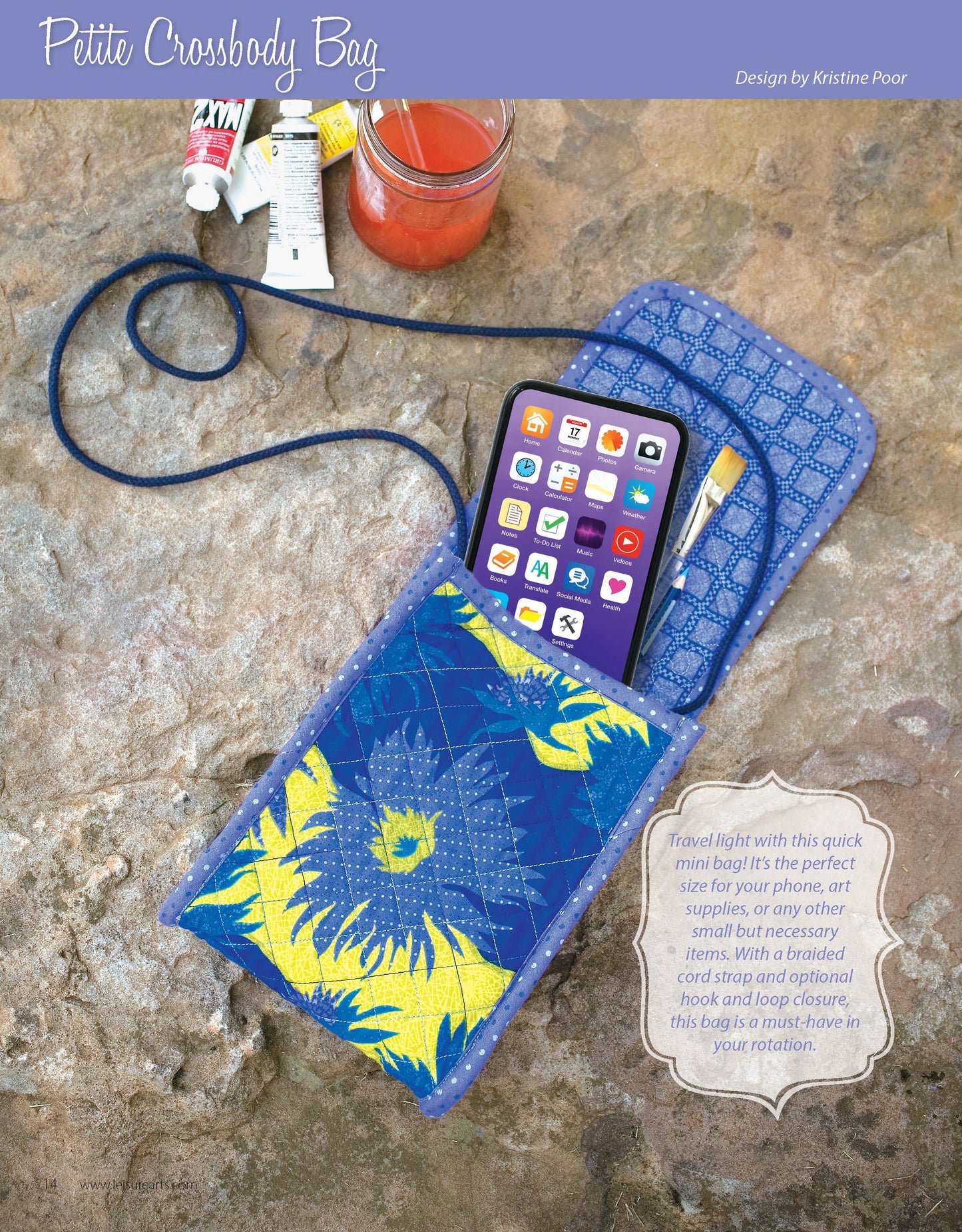 A smartphone rests inside Leisure Arts' "It's in the Bag! Digital Download" quilted crossbody bag, which features a blue body and yellow-blue floral flap. Nearby are paint tubes, brushes, and a drink—perfect for custom bag design inspiration.