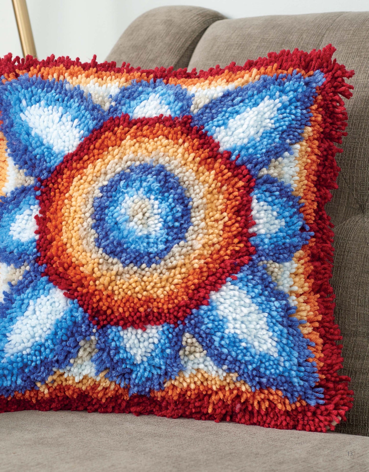 The Timeless Tiles in Latch Hook pillow by Leisure Arts features a vibrant geometric pattern in blue, red, orange, and white. Its textured, fluffy starburst effect stands out atop the brown sofa, inspired by beginner latch hook designs.