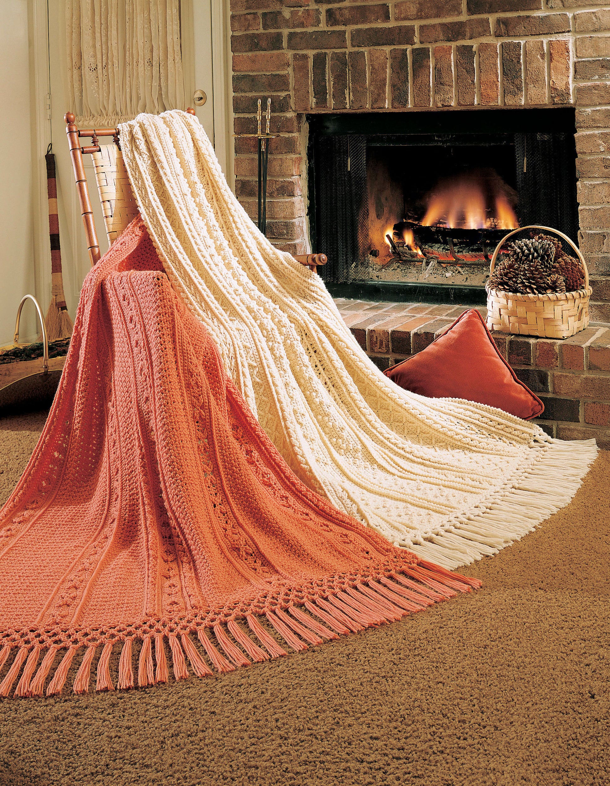 Two medium-weight knit blankets, one peach and one cream, from Fisherman Crochet Afghans by Leisure Arts are draped over a chair near a brick fireplace. A basket of pinecones and a red pillow add to this cozy, rustic scene.