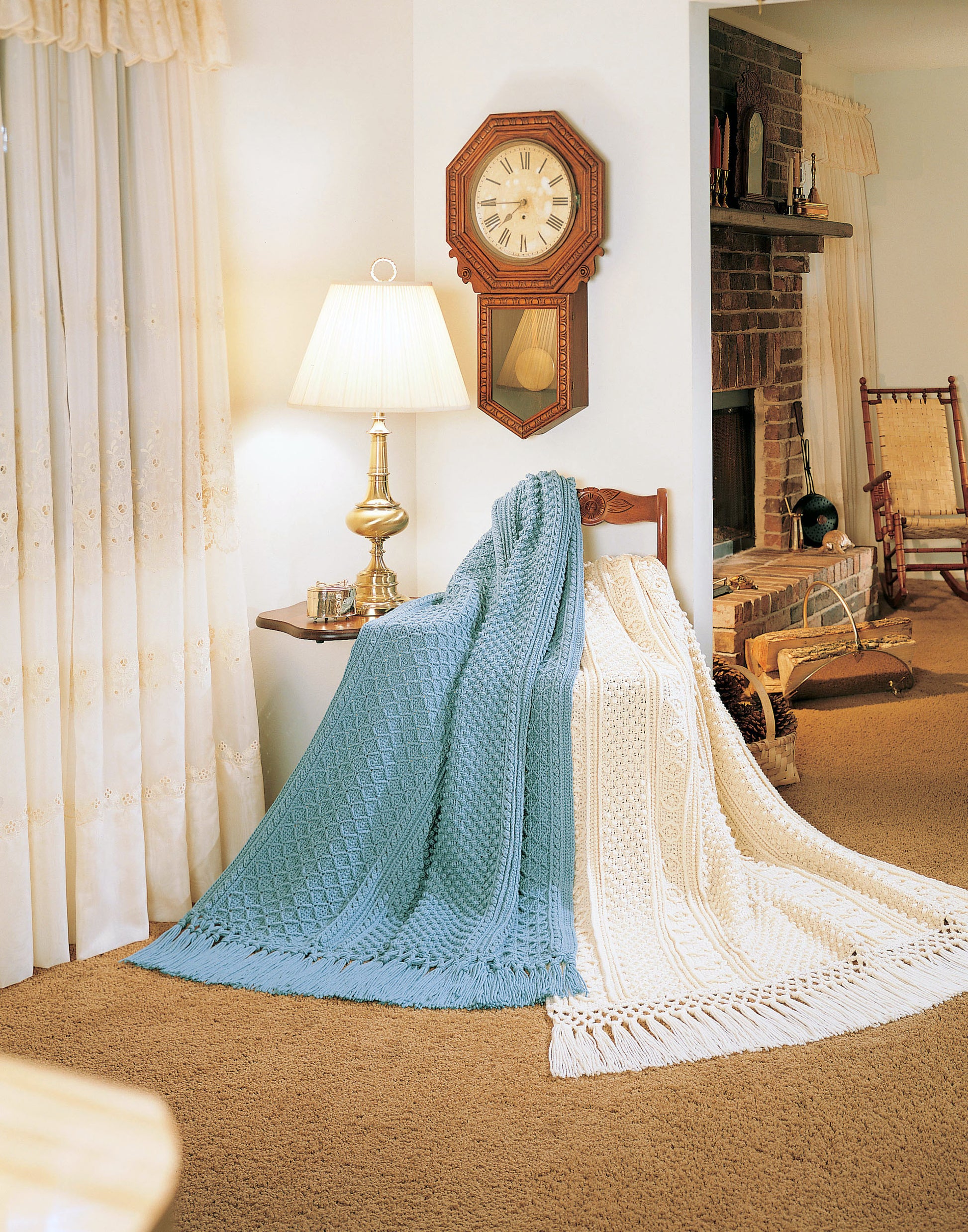 A cozy living room features blue and cream knitted blankets, inspired by Fisherman Crochet Afghans from Leisure Arts, draped over a wooden chair. A lamp, vintage clock, fireplace, beige carpet, and lace curtains complete the warmly lit space.