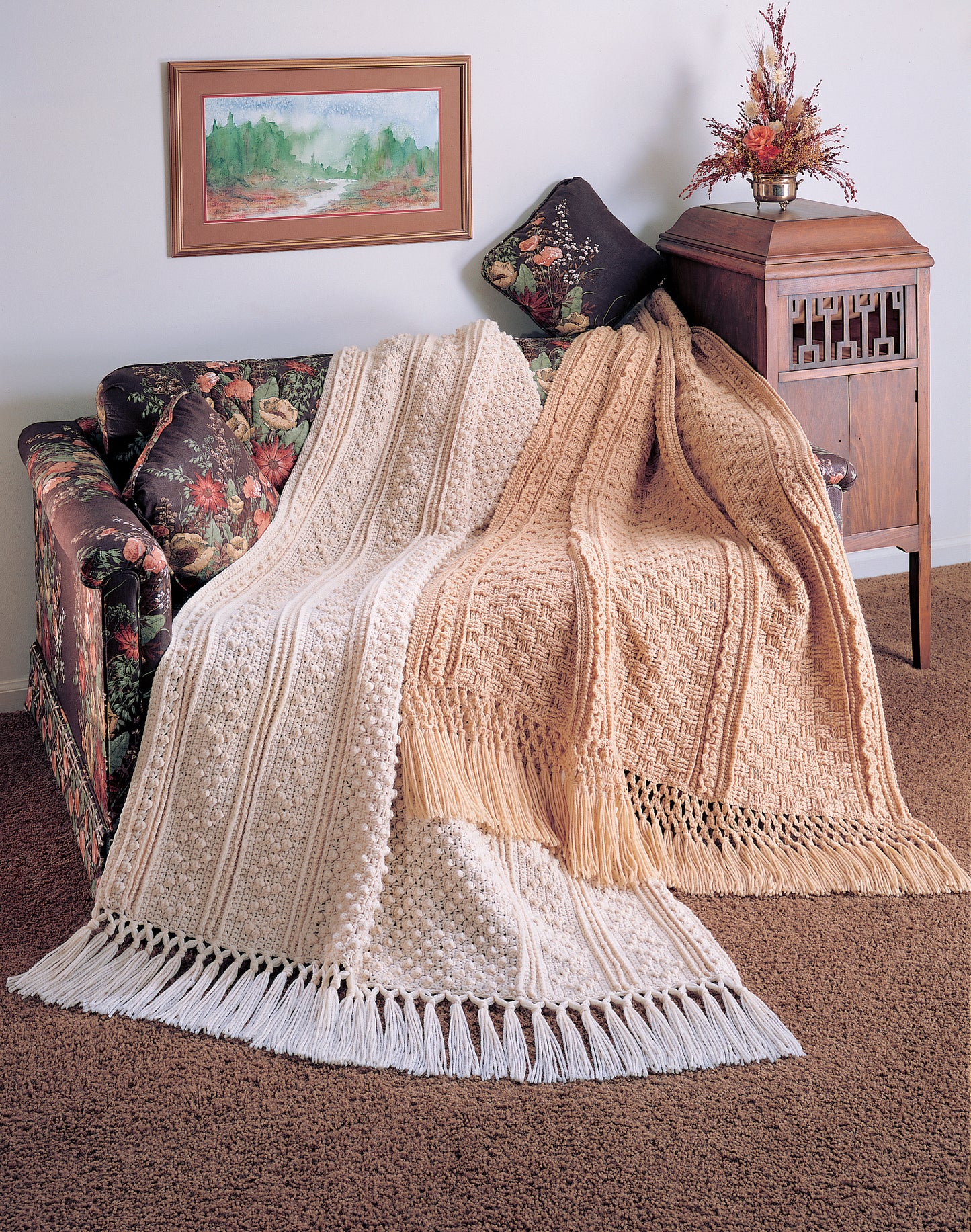Two Leisure Arts Fisherman Crochet Afghans, one cream and one tan with fringe, are draped over a floral-patterned sofa by a vintage wooden cabinet. A framed landscape painting and a floral arrangement decorate the background.