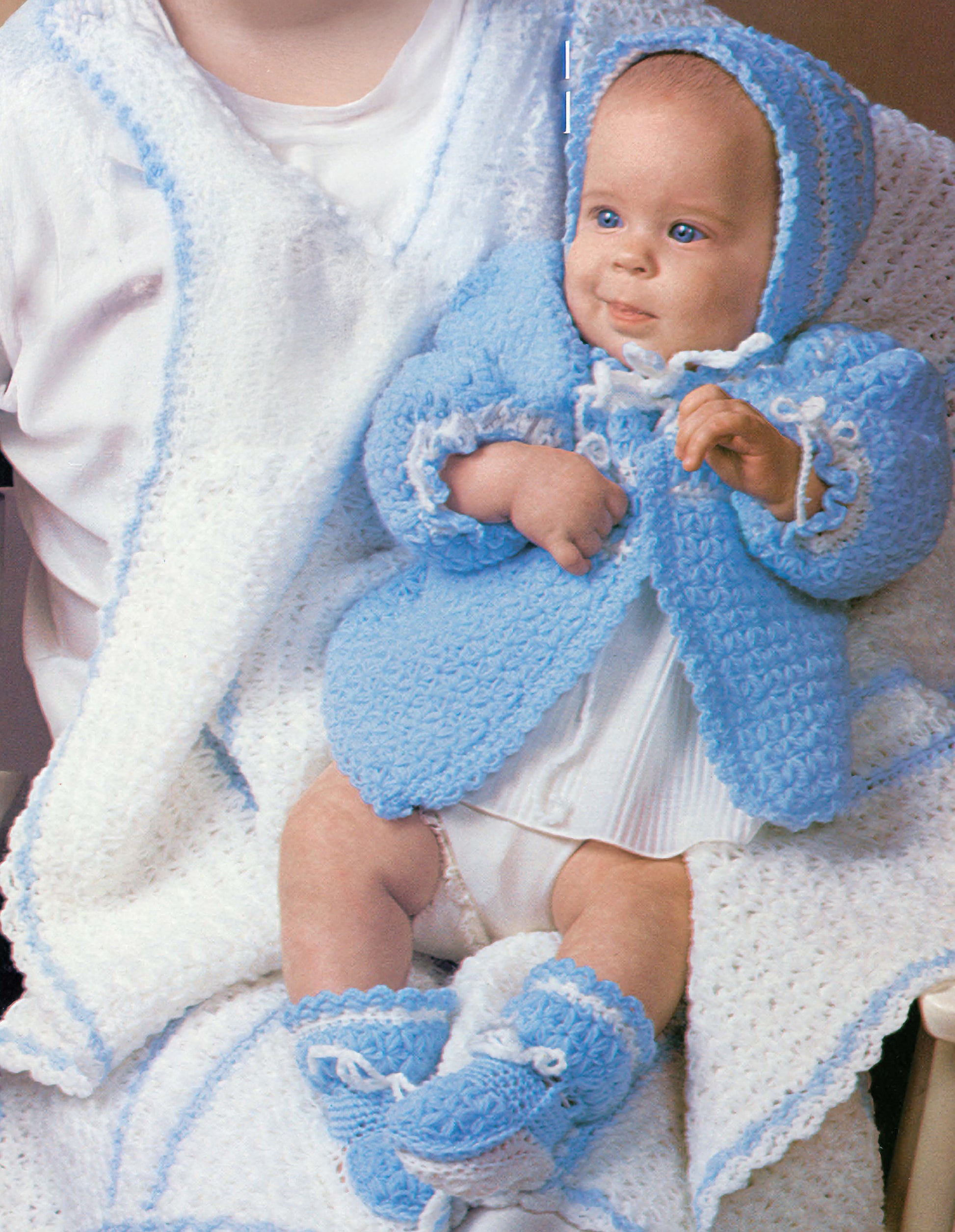 A baby in the Leisure Arts Snowflake Layette to Crochet set—blue bonnet, jacket, and booties—sits on a white crocheted blanket with blue trim and snowflake stitch, held by an adult and gazing up with curiosity.