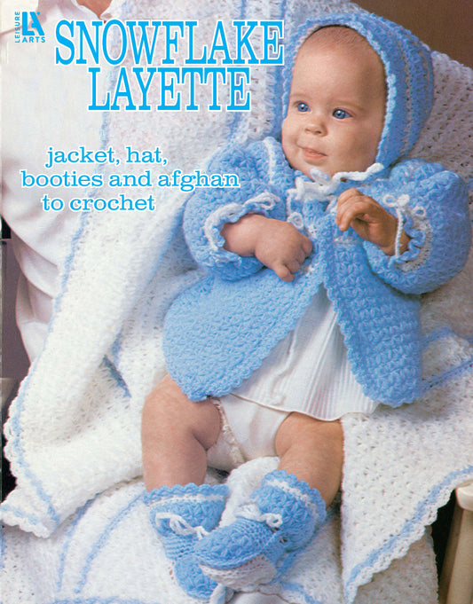 A baby in a blue crocheted jacket, hat, and booties sits on a white afghan. Text: Snowflake Layette to Crochet by Leisure Arts uses snowflake stitch for matching blanket, jacket, hat, and booties.