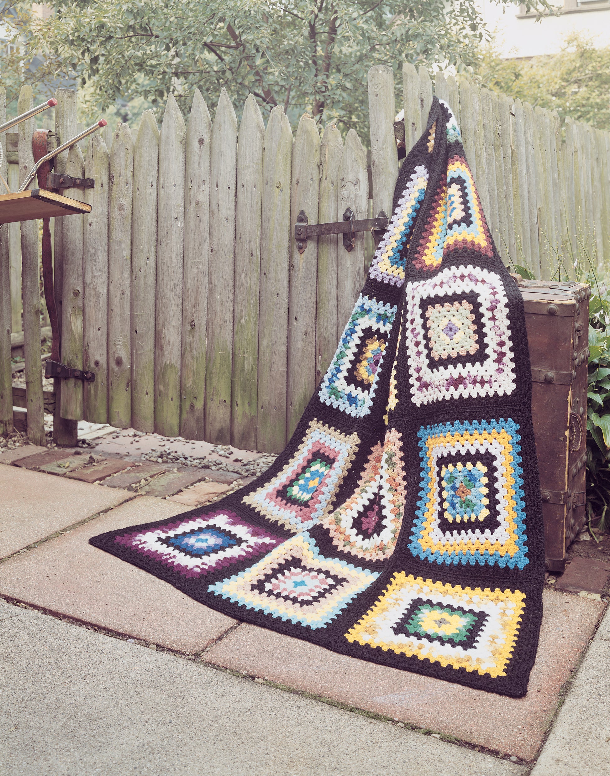 The Afghan Book by Leisure Arts is displayed atop a wooden chest near a weathered picket fence, adding charm to the garden decor.