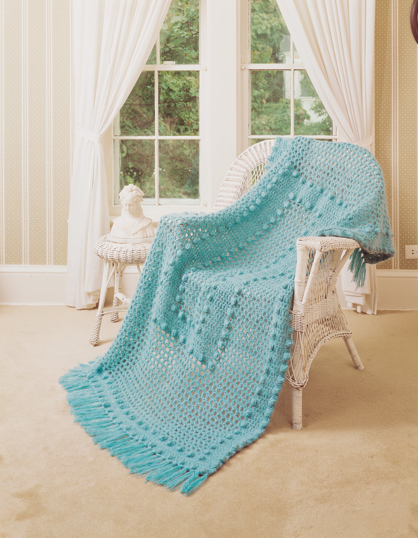 The Afghan Book by Leisure Arts is displayed on a white wicker chair in a bright room with beige carpet, cream wallpaper, and large windows with white curtains.