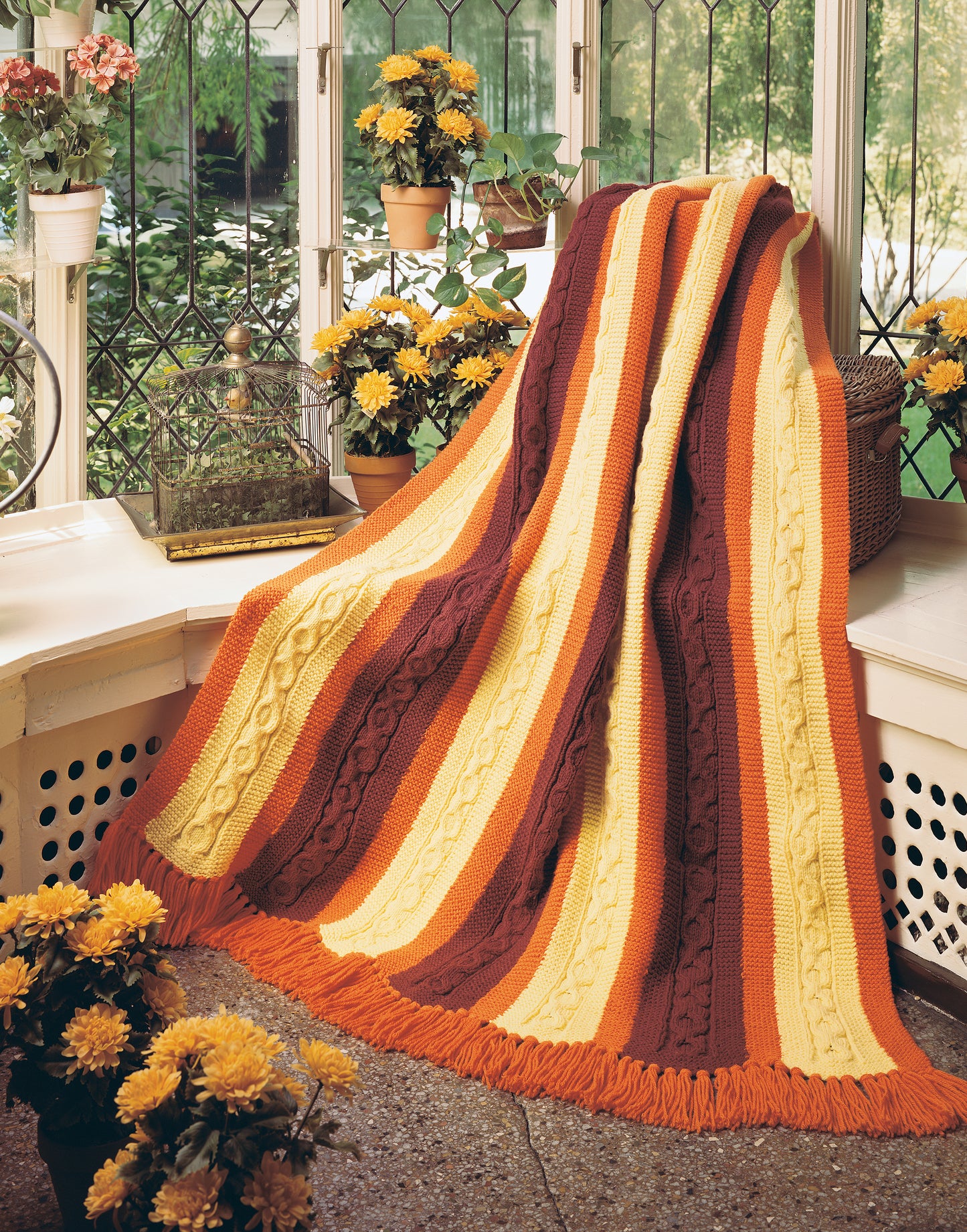 The Afghan Book by Leisure Arts features a cozy knitted blanket with wide orange, yellow, and brown stripes draped over a white bench surrounded by yellow flowers in a sunlit room.