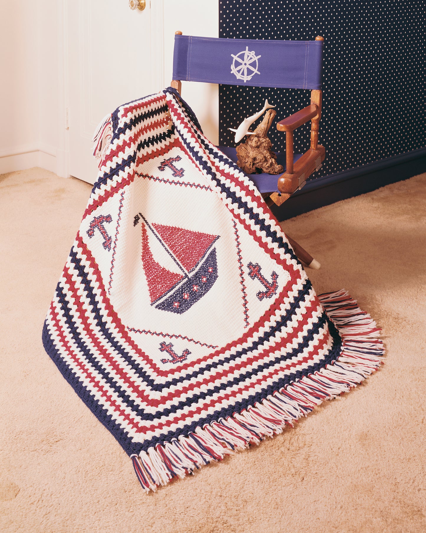 The Afghan Book by Leisure Arts is featured draped over a wooden chair with a blue nautical backrest, on beige carpet in a blue and white room. The crocheted blanket has red sailboats and blue anchors in a patchwork afghan style.