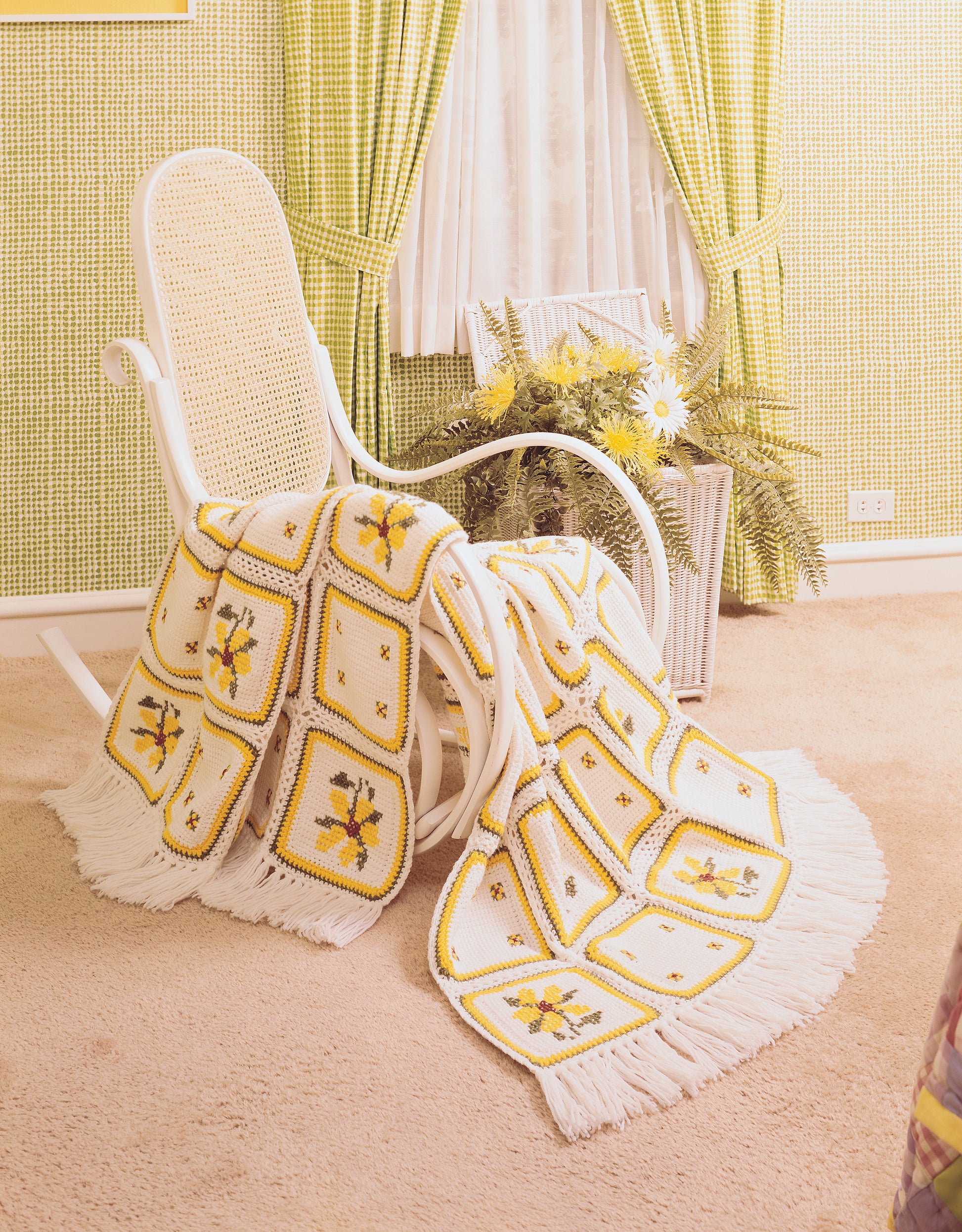 A white rocking chair holds a fringed patchwork afghan made using patterns from The Afghan Book by Leisure Arts, with yellow and brown flowers. It sits on beige carpet near a basket of yellow and white flowers in a retro-styled room.