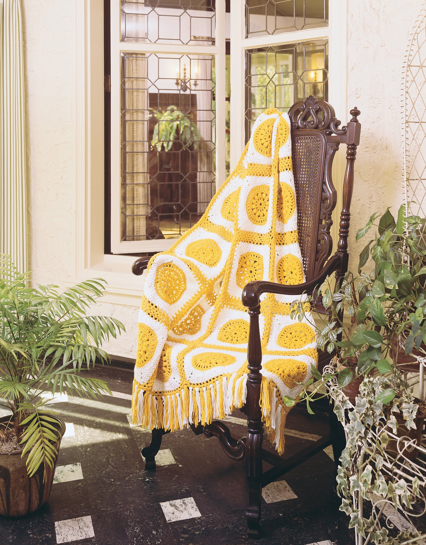 The Afghan Book by Leisure Arts is displayed on an ornate dark wooden chair draped with a yellow and white crocheted blanket, surrounded by green plants in front of a large window with decorative glass panes.