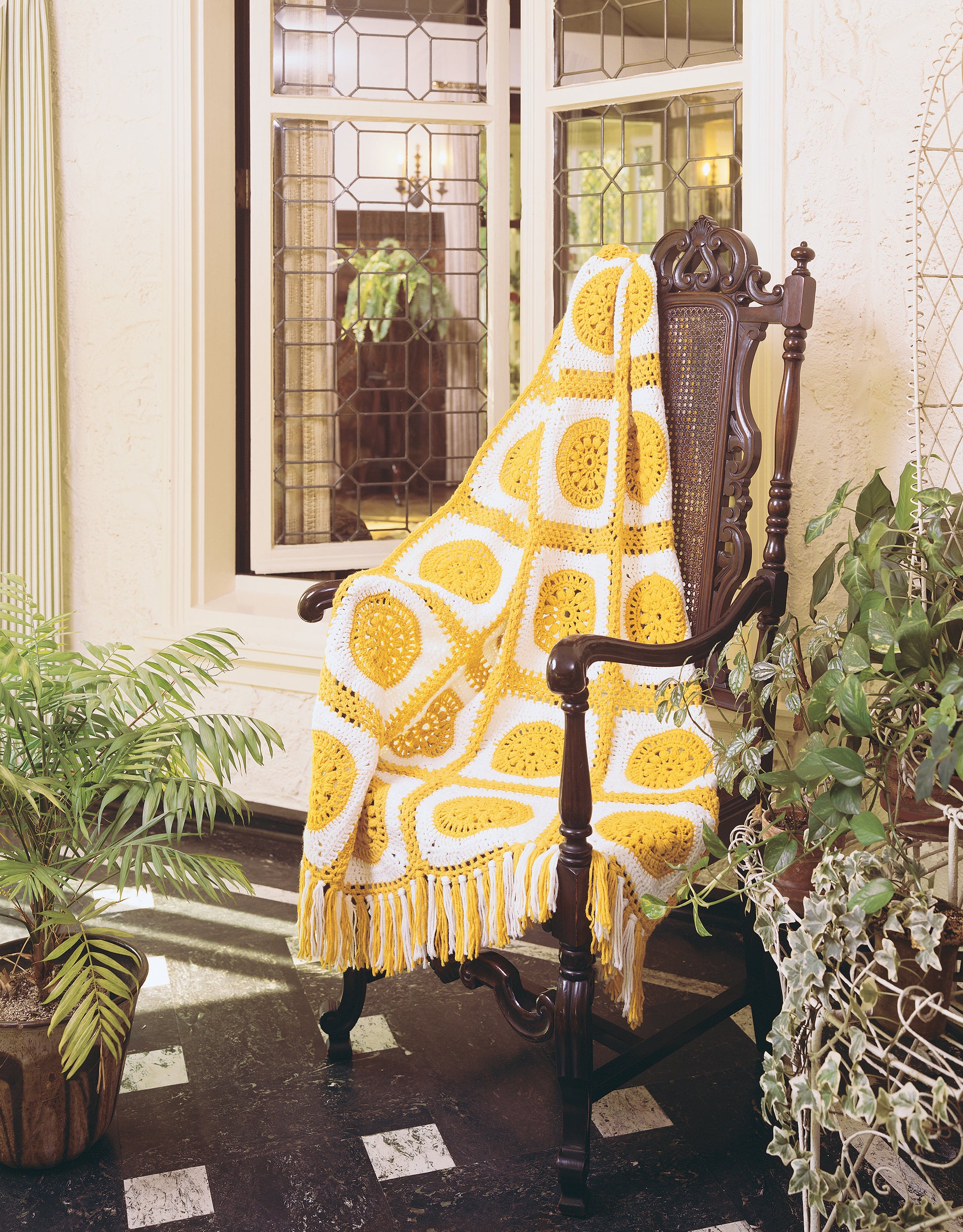 The Afghan Book by Leisure Arts is displayed on an ornate dark wooden chair draped with a yellow and white crocheted blanket, surrounded by green plants in front of a large window with decorative glass panes.