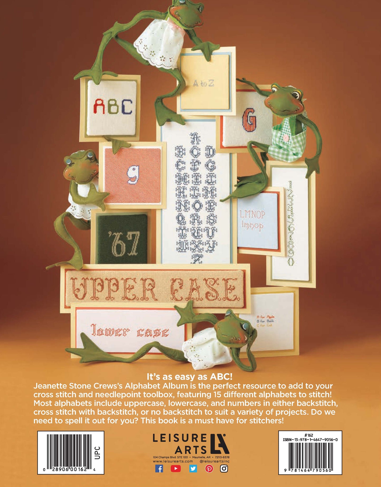 Four plush frogs climb among framed cross stitch alphabets, displayed on an orange background. At the bottom, text describes "Alphabet Album—15 Decorative Alphabets for Cross Stitch and Needlepoint" by Leisure Arts. A barcode appears at lower right.