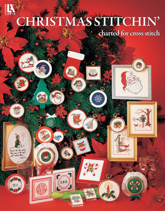 A Christmas tree is adorned with beginner-friendly cross-stitched ornaments from Leisure Arts' Christmas Stitchin', featuring festive designs like Santa, wreaths, stockings, and reindeer. Framed cross-stitch art decorates the base.